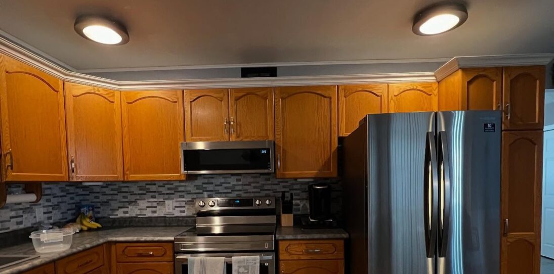 Dated L-shaped kitchen with honey orange oak cathedral-arch upper cabinets, white crown molding running along the ceiling, a gray and white glass mosaic backsplash, dark speckled counters, a stainless steel electric range with an over-the-range microwave, a tall stainless French-door fridge on the right, and two bronze flush-mount ceiling lights.
