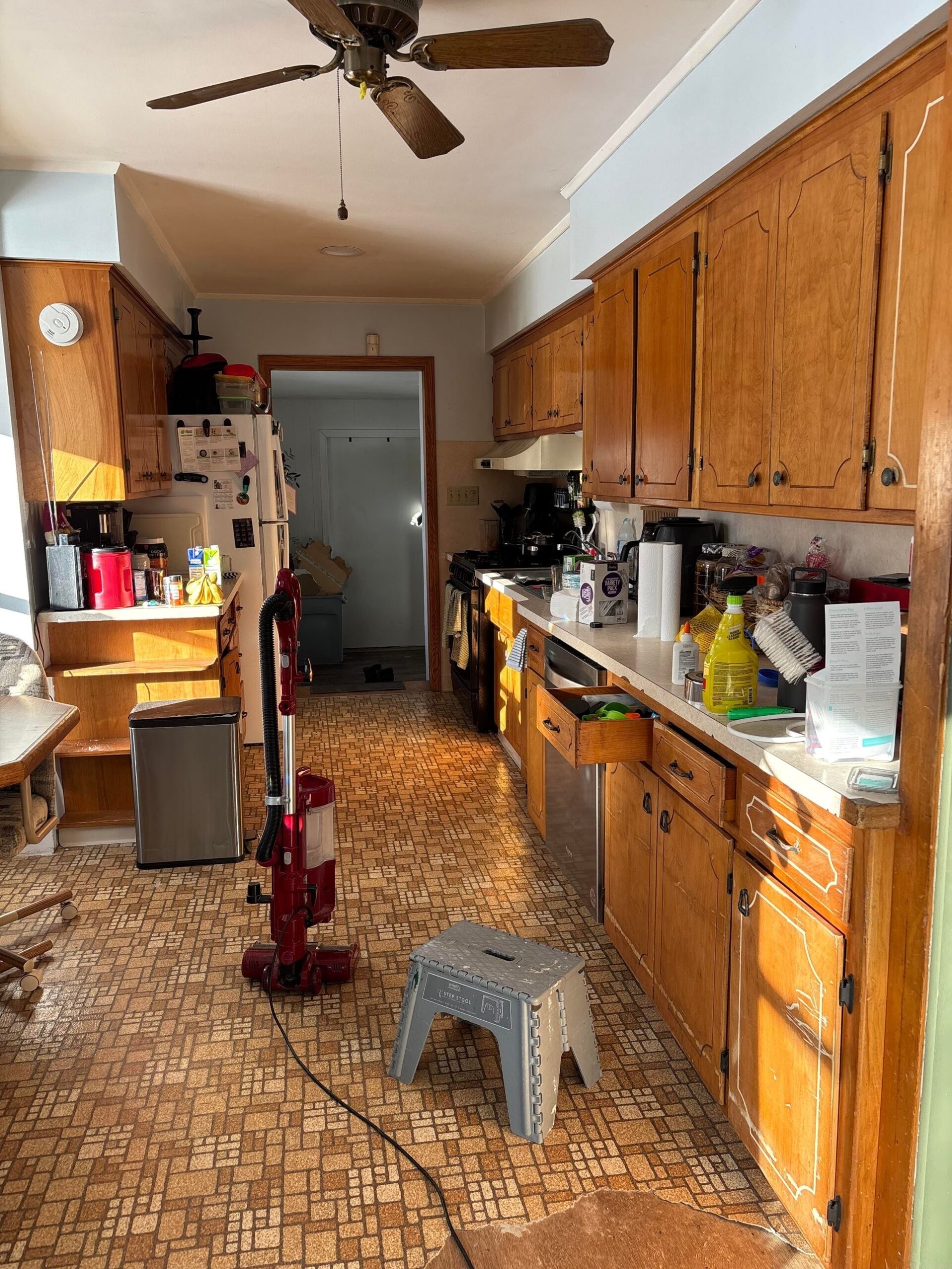 Narrow galley kitchen with warm orange-toned oak cabinets, brown geometric linoleum floor, a dark wood ceiling fan, a red upright vacuum and a gray step stool on the floor, and a stainless step-can near the fridge.