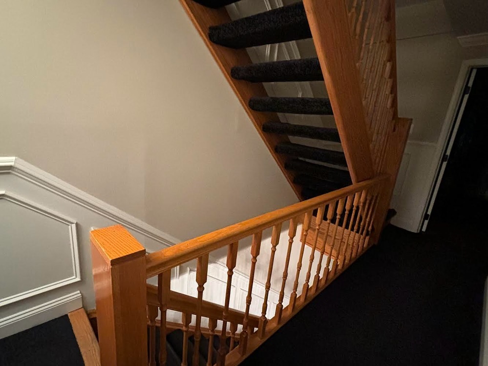 Upstairs landing with an orange oak railing, matching spindles and newel post, dark carpet runner on the stair treads and white panel molding on the far left wall.