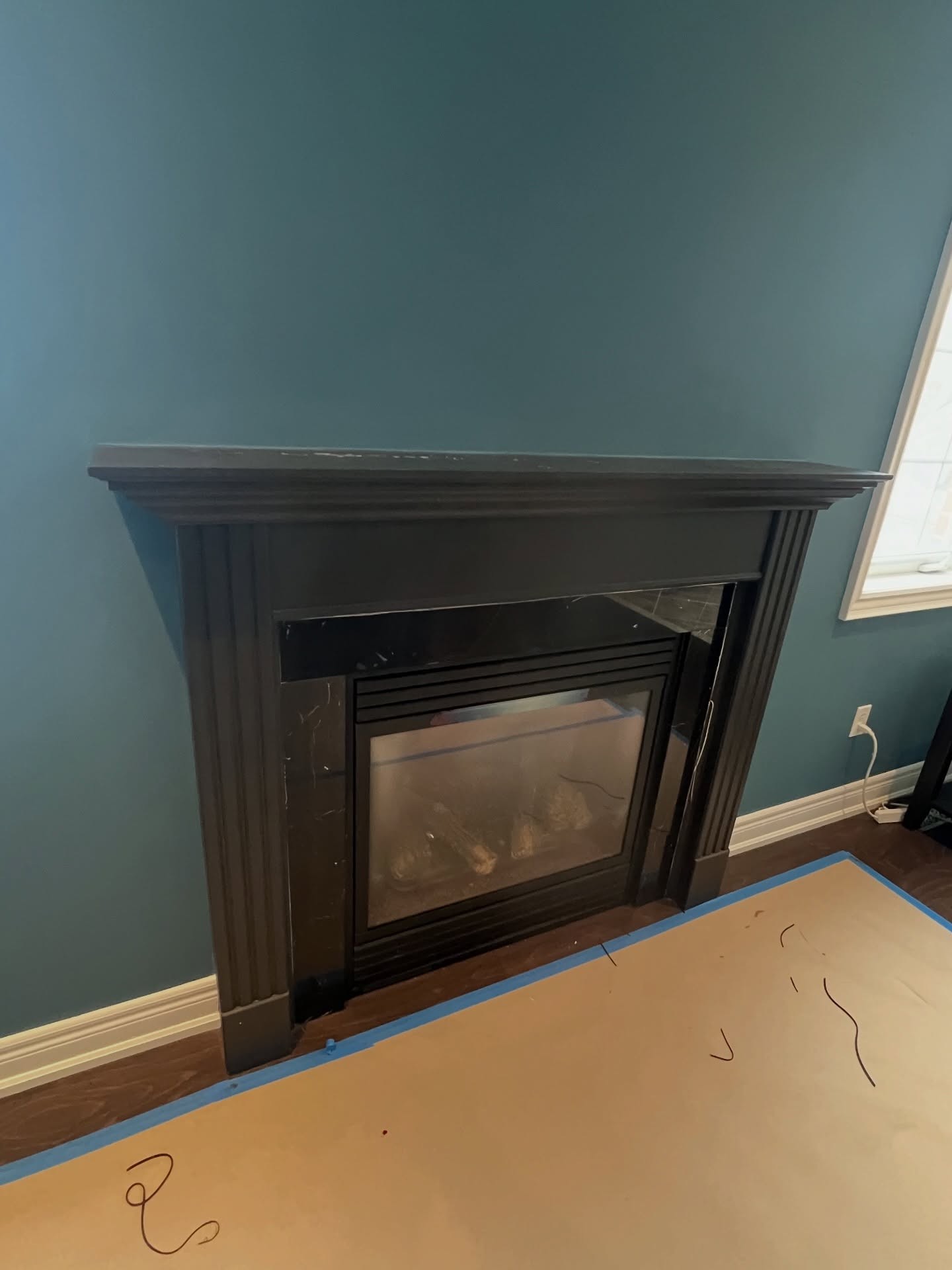 Dark espresso wood mantel with carved pilasters, dark marble inset, and a black gas insert, mounted against a dark teal wall with a long stretch of empty blue wall above it.