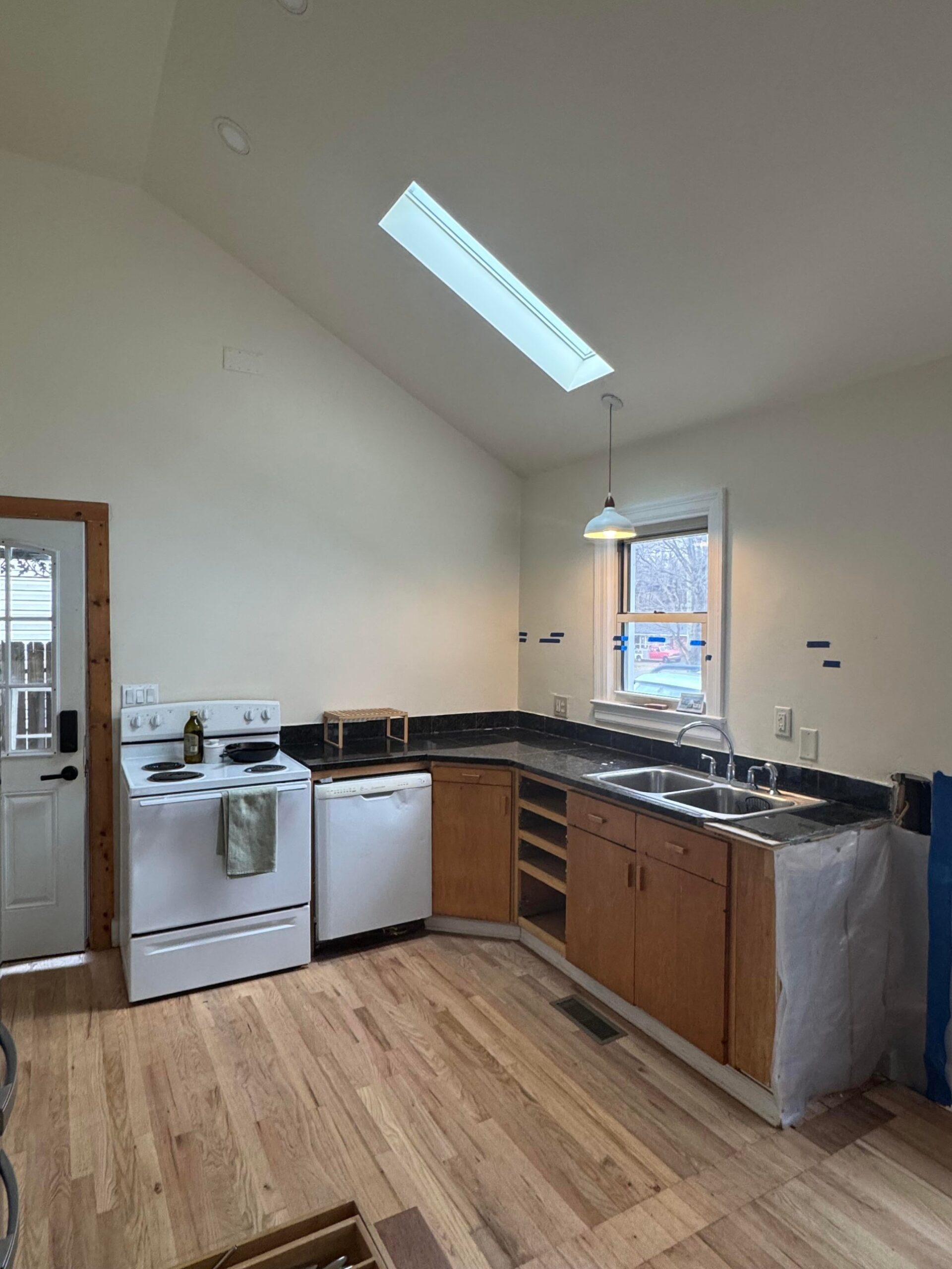 Small sloped-ceiling kitchen with a white electric range, white dishwasher, basic brown builder-grade lower cabinets, dark tile counters, and a double stainless sink under the window.