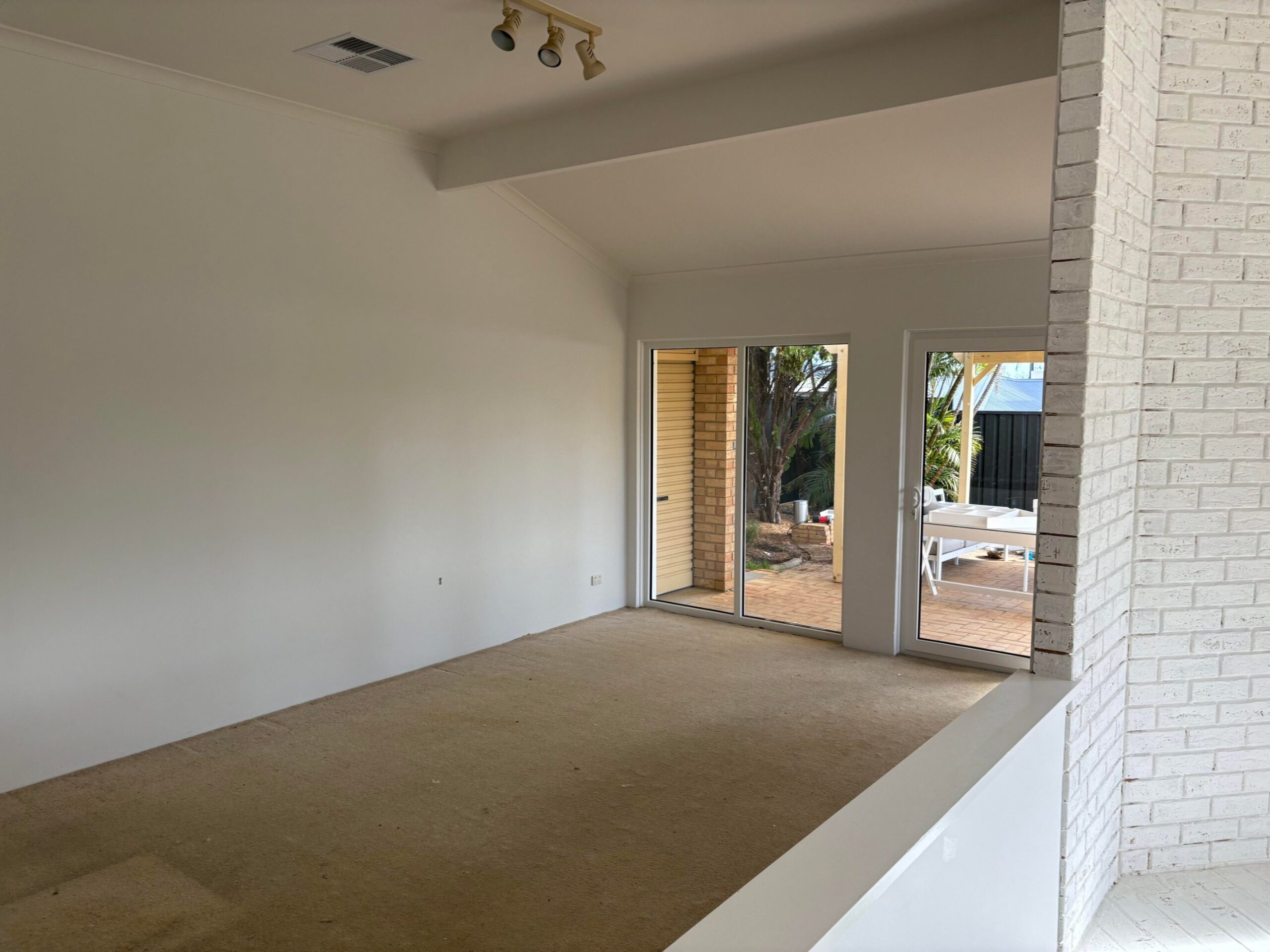 Empty living room with beige carpet, white painted brick accent, sliding doors to a patio, and vaulted ceilings with a wood beam.