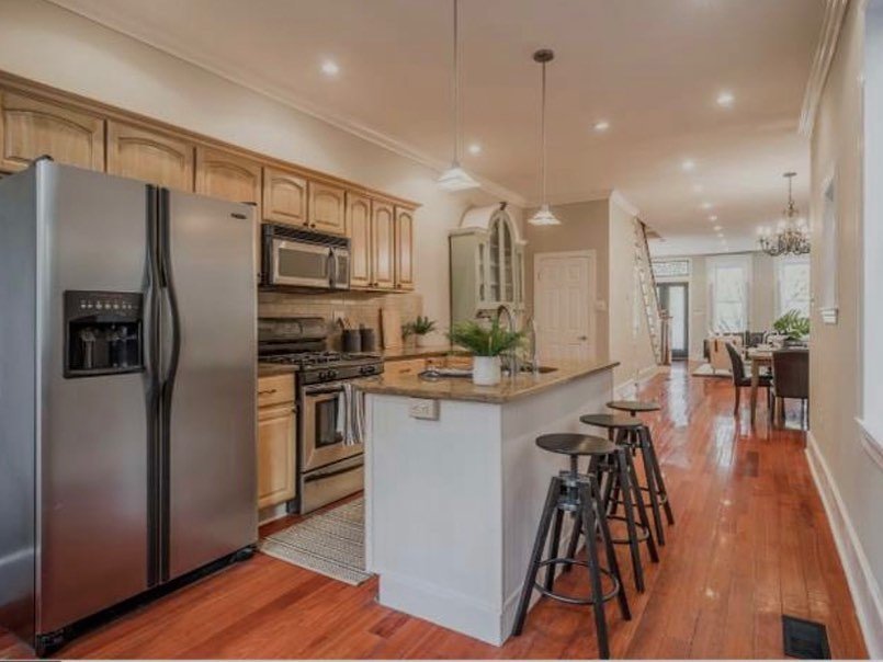 Wide view of the original kitchen with honey oak upper and lower cabinets running down a galley layout, cherry hardwood floors, a white beadboard island with gold granite top, pendant lights overhead, and warm neutral walls.