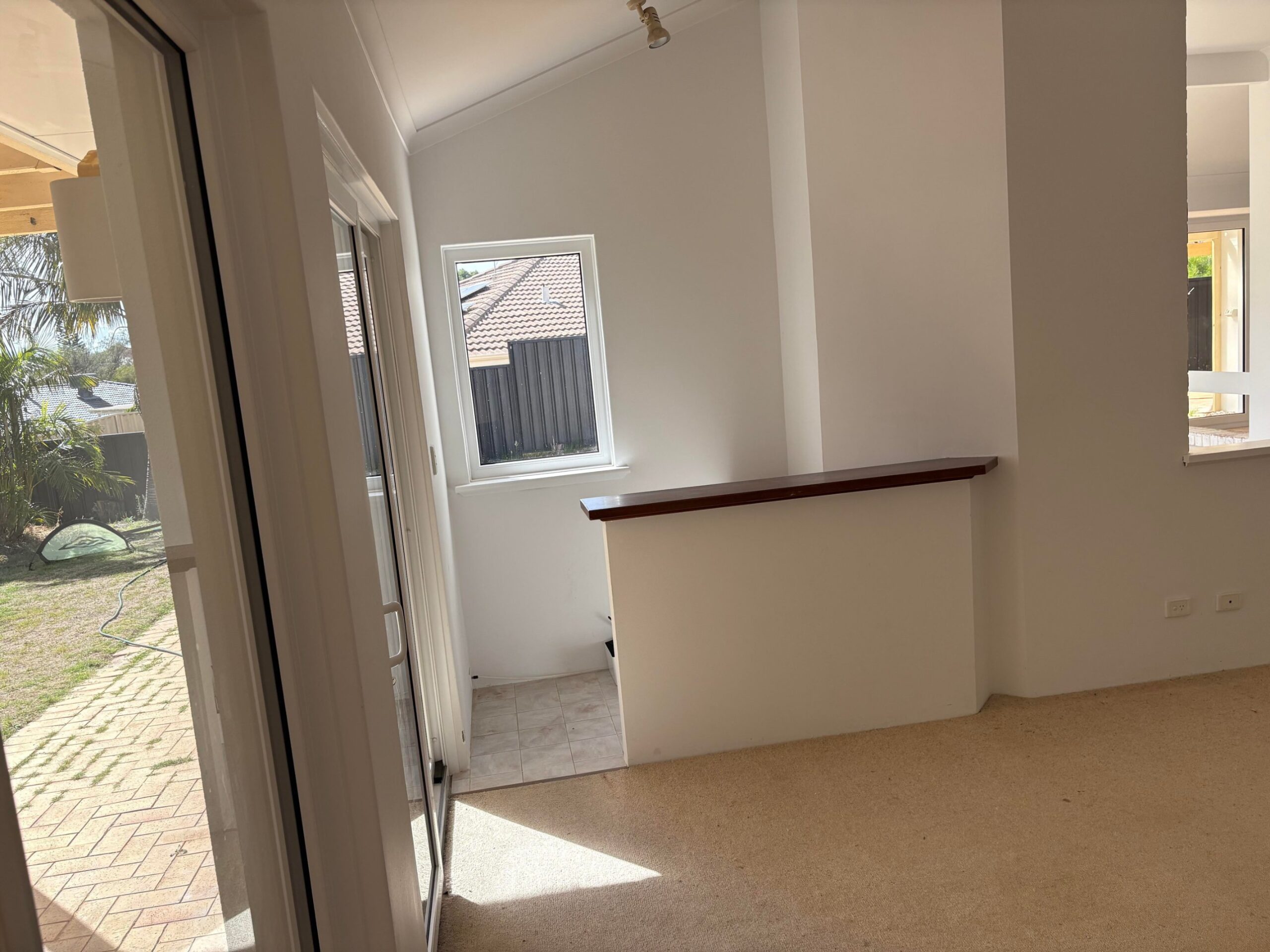Dated sunroom corner with beige carpet, white painted brick walls, a dark wood ledge, and a small window with sliding door view.