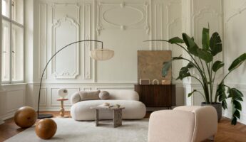 Parisian-style living room with ornate white plaster wall paneling and crown molding, a curved cream boucle sofa paired with a matching boxy lounge chair, a black arc floor lamp with a pleated silk shade, a dark wood ribbed sideboard, two large wood sphere sculptures resting on an ivory rug, a small sculptural wood coffee table, and a tall bird of paradise plant on a chevron herringbone wood parquet floor.