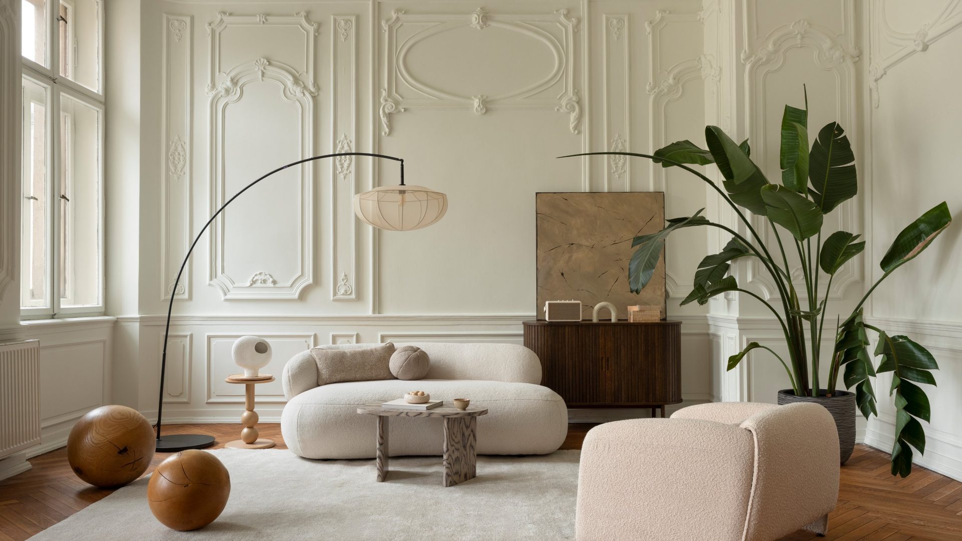 Parisian-style living room with ornate white plaster wall paneling and crown molding, a curved cream boucle sofa paired with a matching boxy lounge chair, a black arc floor lamp with a pleated silk shade, a dark wood ribbed sideboard, two large wood sphere sculptures resting on an ivory rug, a small sculptural wood coffee table, and a tall bird of paradise plant on a chevron herringbone wood parquet floor.