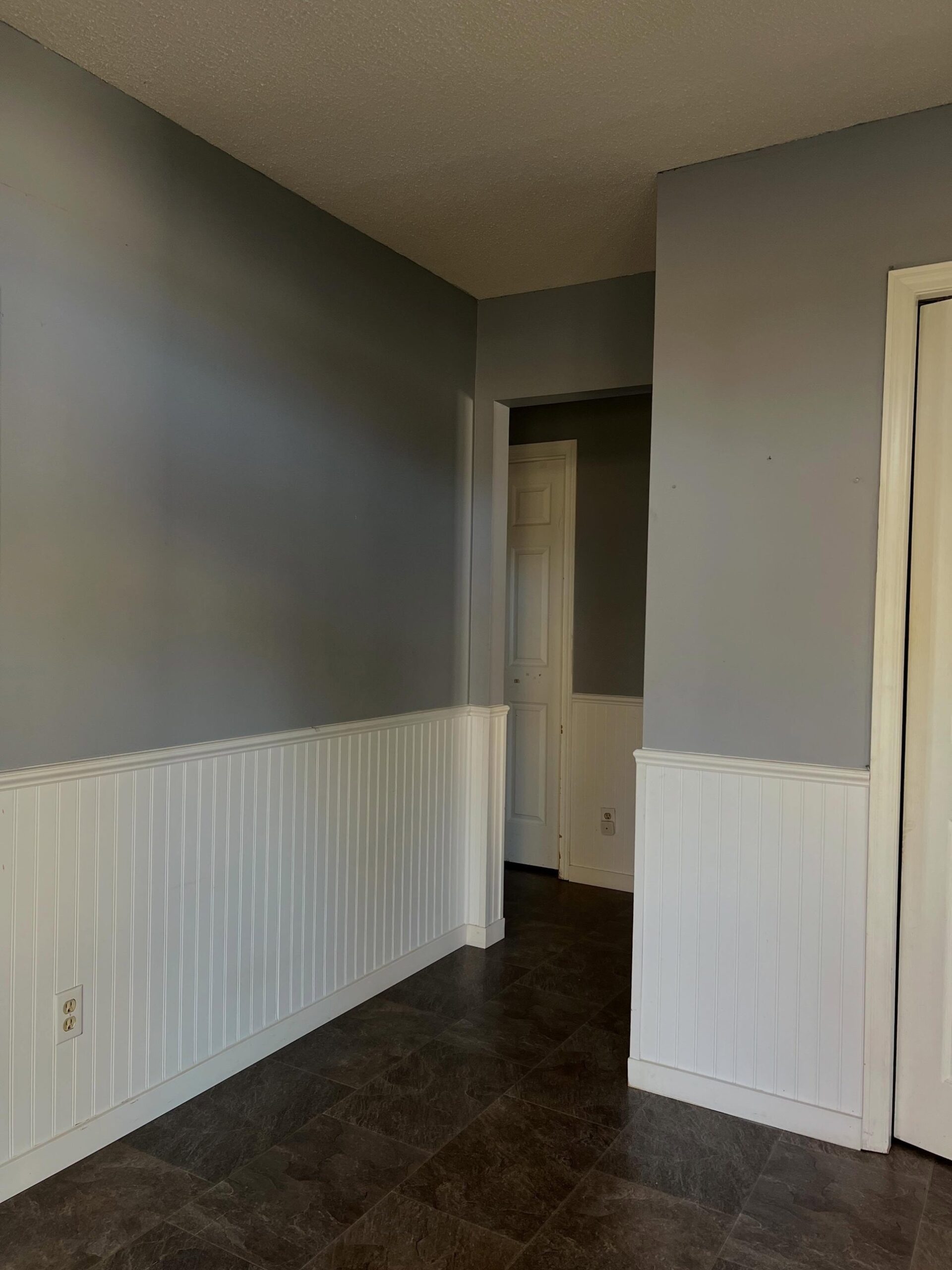 Corner view of the same space in its original pigeon-gray paint, showing a wall section, a doorway opening, white beadboard wainscoting, and a dark slate tile floor.