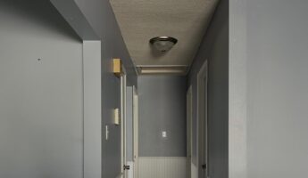 Narrow upstairs hallway with soft pigeon-gray upper walls, white beadboard wainscoting below, textured popcorn ceiling with a brushed-nickel flush mount light centered above, white doors on either side, and a dark slate-look tile floor stretching down the corridor.
