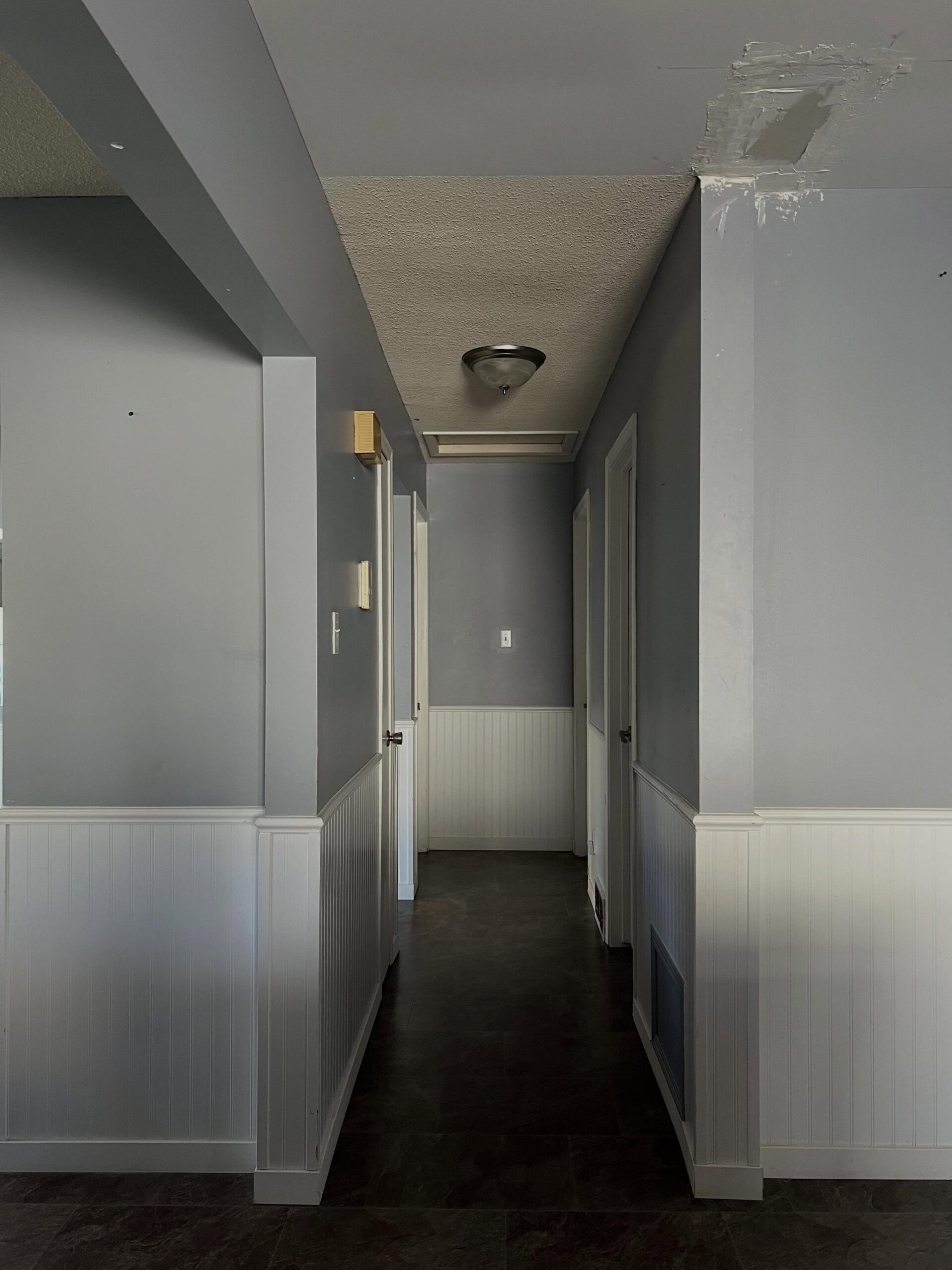 Narrow upstairs hallway painted a soft pigeon-gray on the upper walls with white beadboard wainscoting below, a dark slate-look tile floor, textured ceiling, and a brushed-nickel flush mount light, with a mid-renovation drywall patch visible in the top right corner.