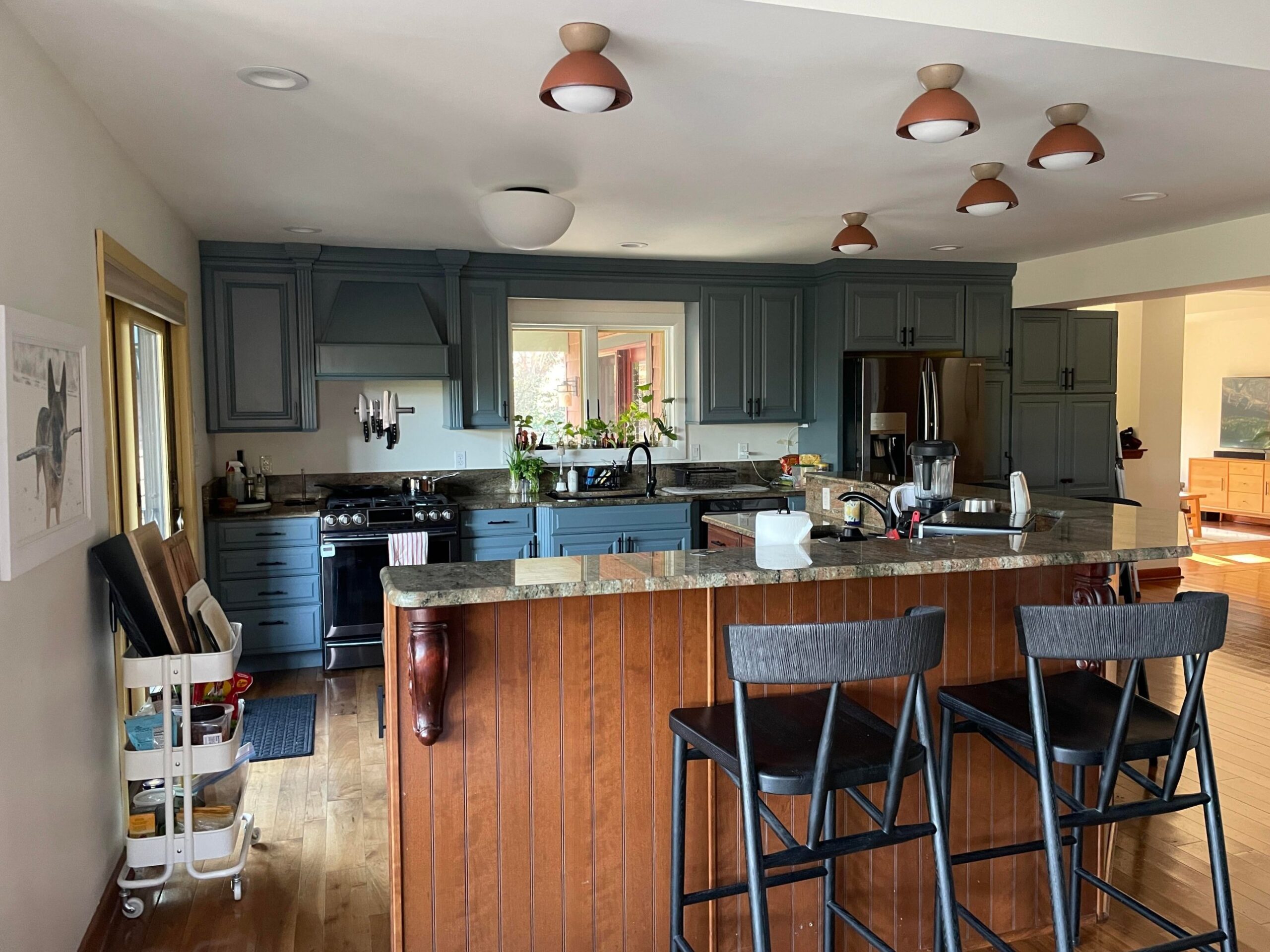 Wide view of the painted blue kitchen with the original brown cherry beadboard island still in place, now reading as an intentional warm wood accent under small terracotta dome ceiling lights.