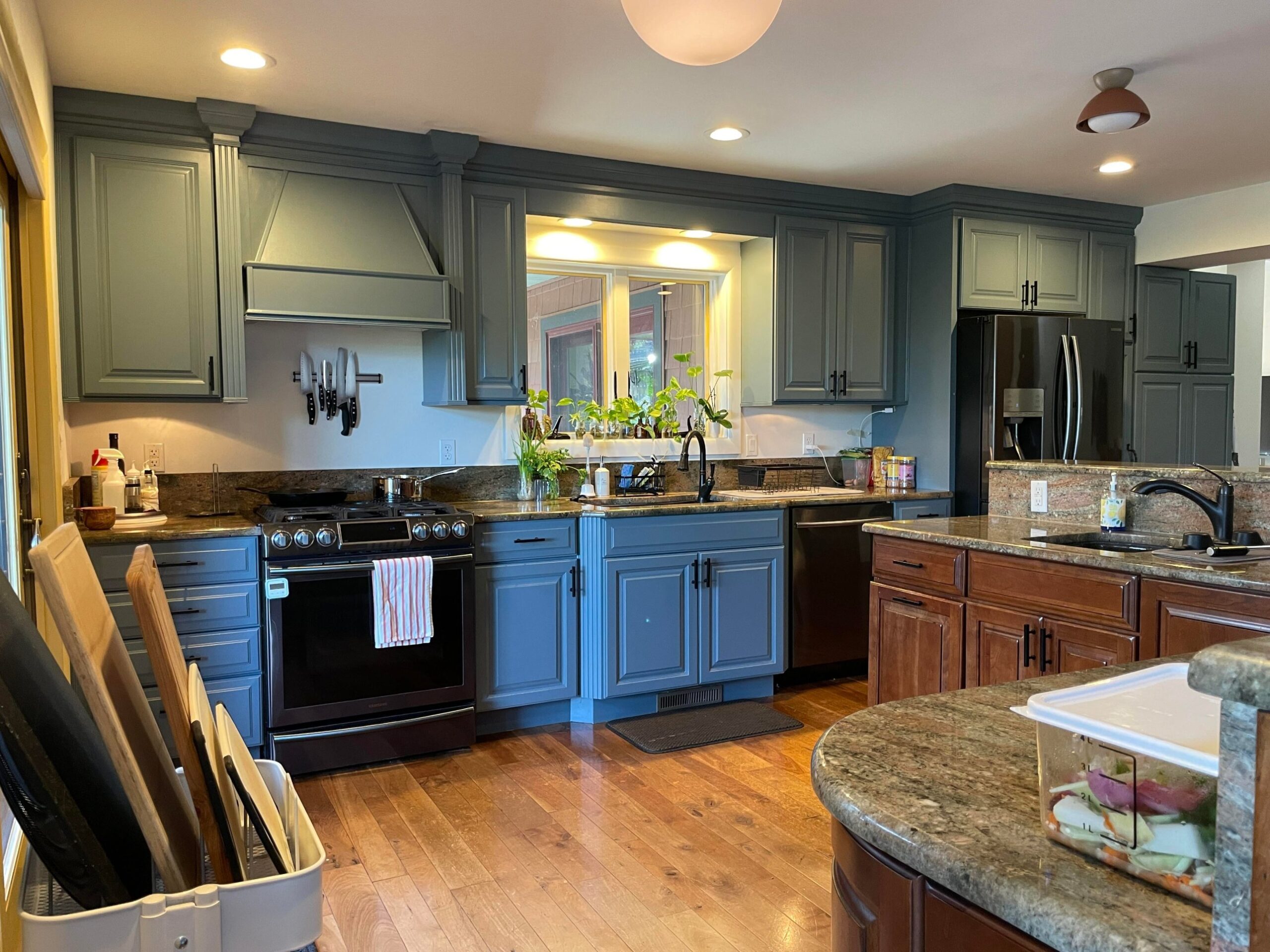 Deep slate blue cabinets around the same black gas range, with the magnetic knife strip, ornate hood, and window now reading as moody traditional under warm terracotta ceiling lights.