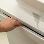 Hand pushing on the top edge of a push-to-open bathroom vanity drawer with a glossy white and marble-patterned front, mounted beneath a white ceramic vessel sink with a chrome drain.