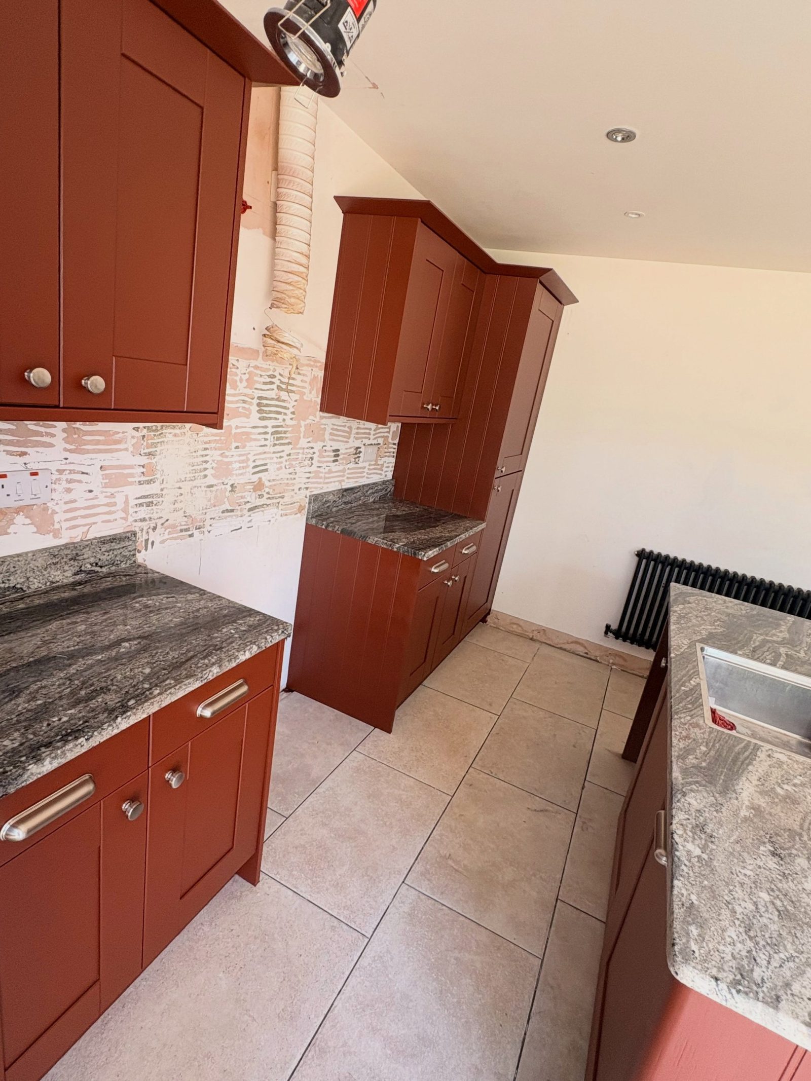 Narrow galley view of the red kitchen with lower cabinets, granite worktops, and a tall red pantry unit at the end.