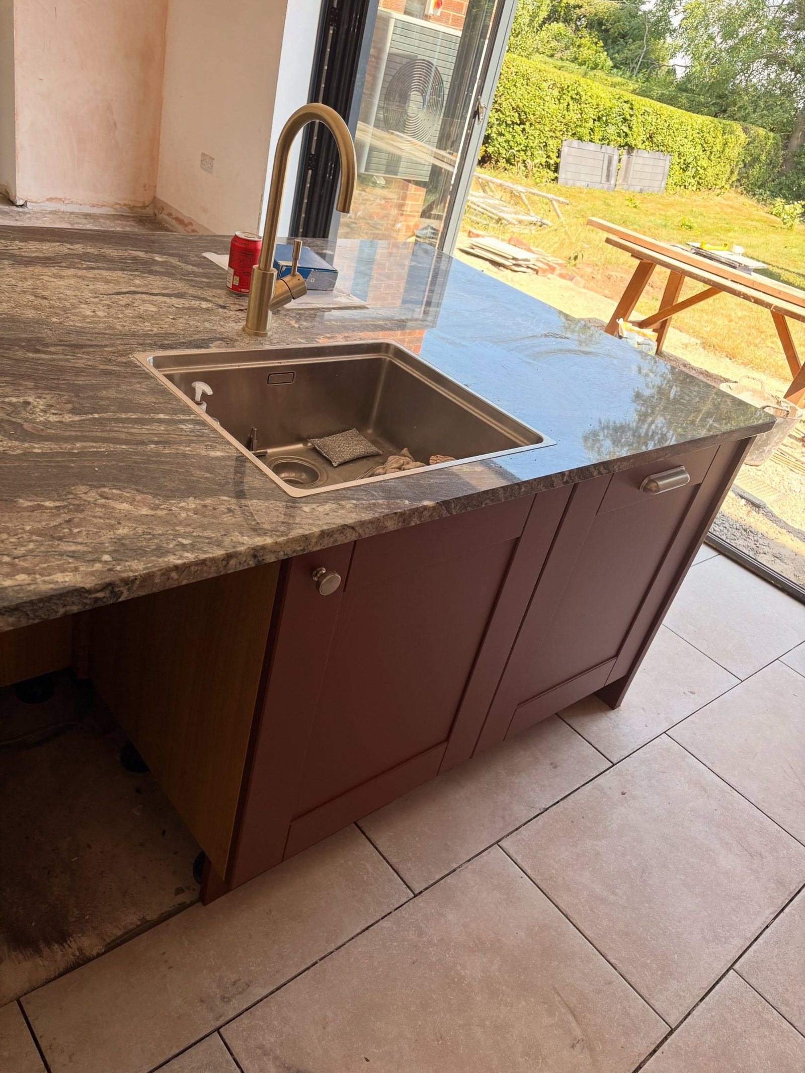 Red kitchen island with inset sink and brass tap, bifold doors fully open to the garden on a sunny day.