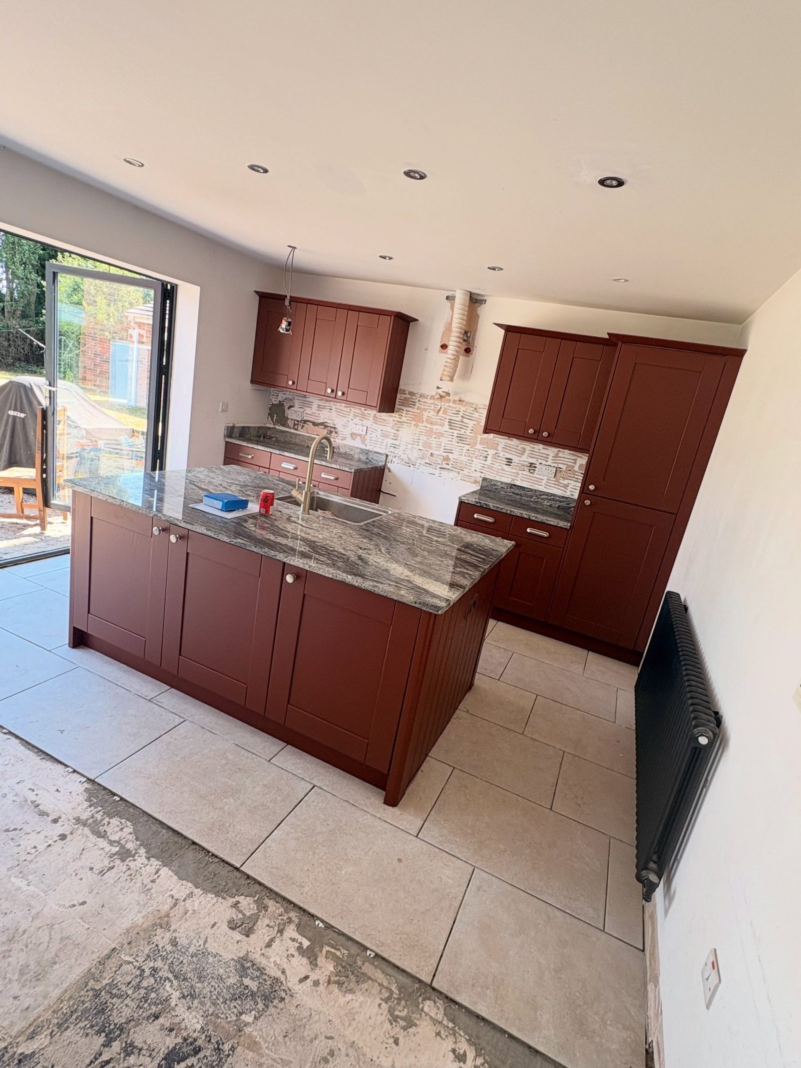 Wide shot of the finished kitchen with Etruscan Red shaker cabinets, central island, column radiator, and bifold doors to the garden.