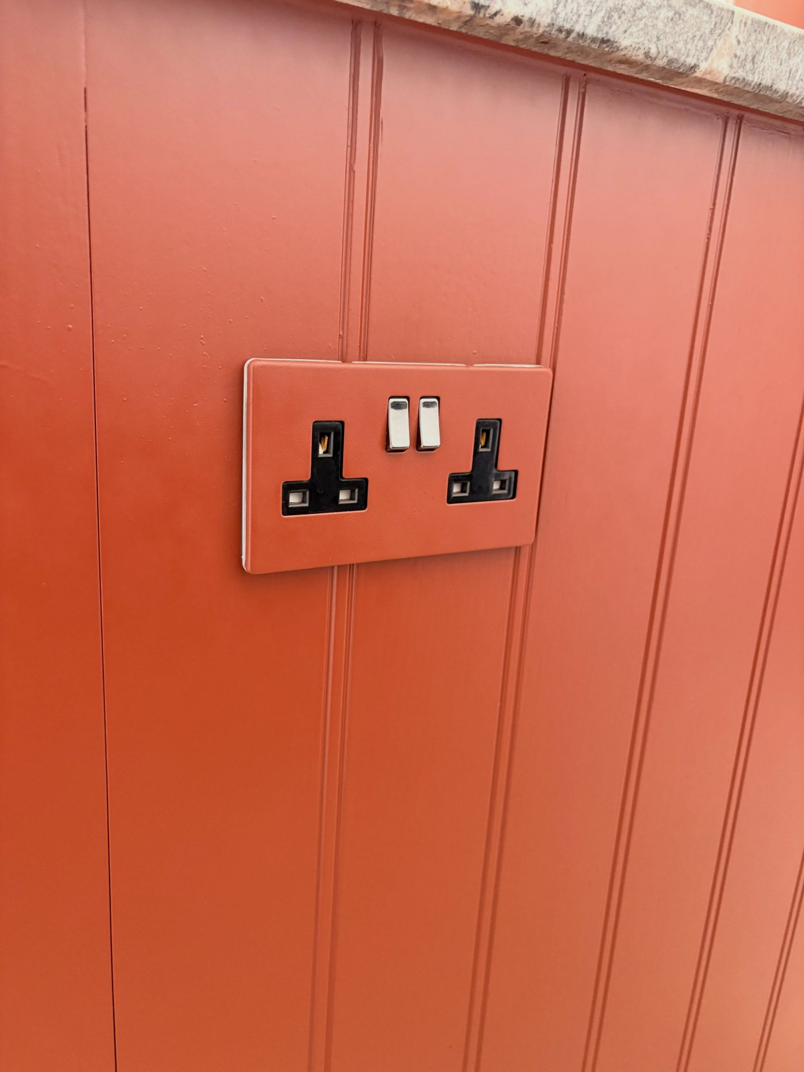 Double UK plug socket painted the same Etruscan Red as the beadboard cabinet end panel it sits on.