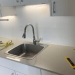 Rental kitchen sink area before the backsplash install, with white flat-front cabinets, a stainless steel drop-in sink, gray quartz counter, and a plain blank white painted wall behind the sink where the green peel-and-stick subway tiles will later go.