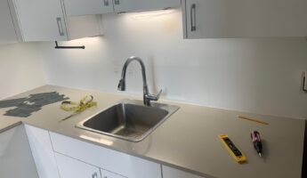 Rental kitchen sink area before the backsplash install, with white flat-front cabinets, a stainless steel drop-in sink, gray quartz counter, and a plain blank white painted wall behind the sink where the green peel-and-stick subway tiles will later go.
