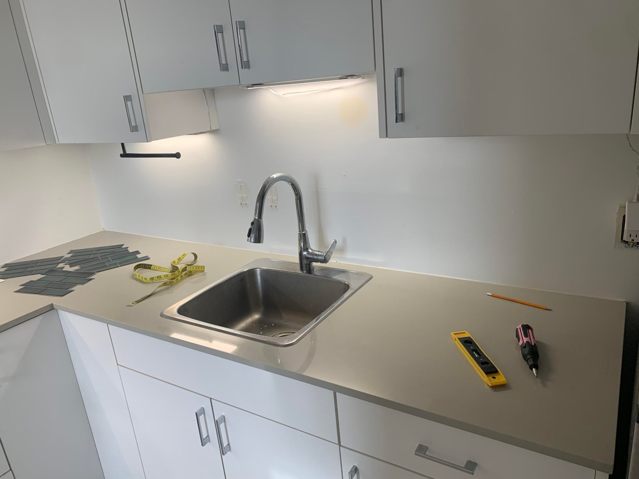 Rental kitchen with white flat-front cabinets, a stainless steel drop-in sink, a plain painted white backsplash, and a stack of green peel-and-stick subway tiles on the counter next to a yellow tape measure, a yellow bubble level, and a pink screwdriver.