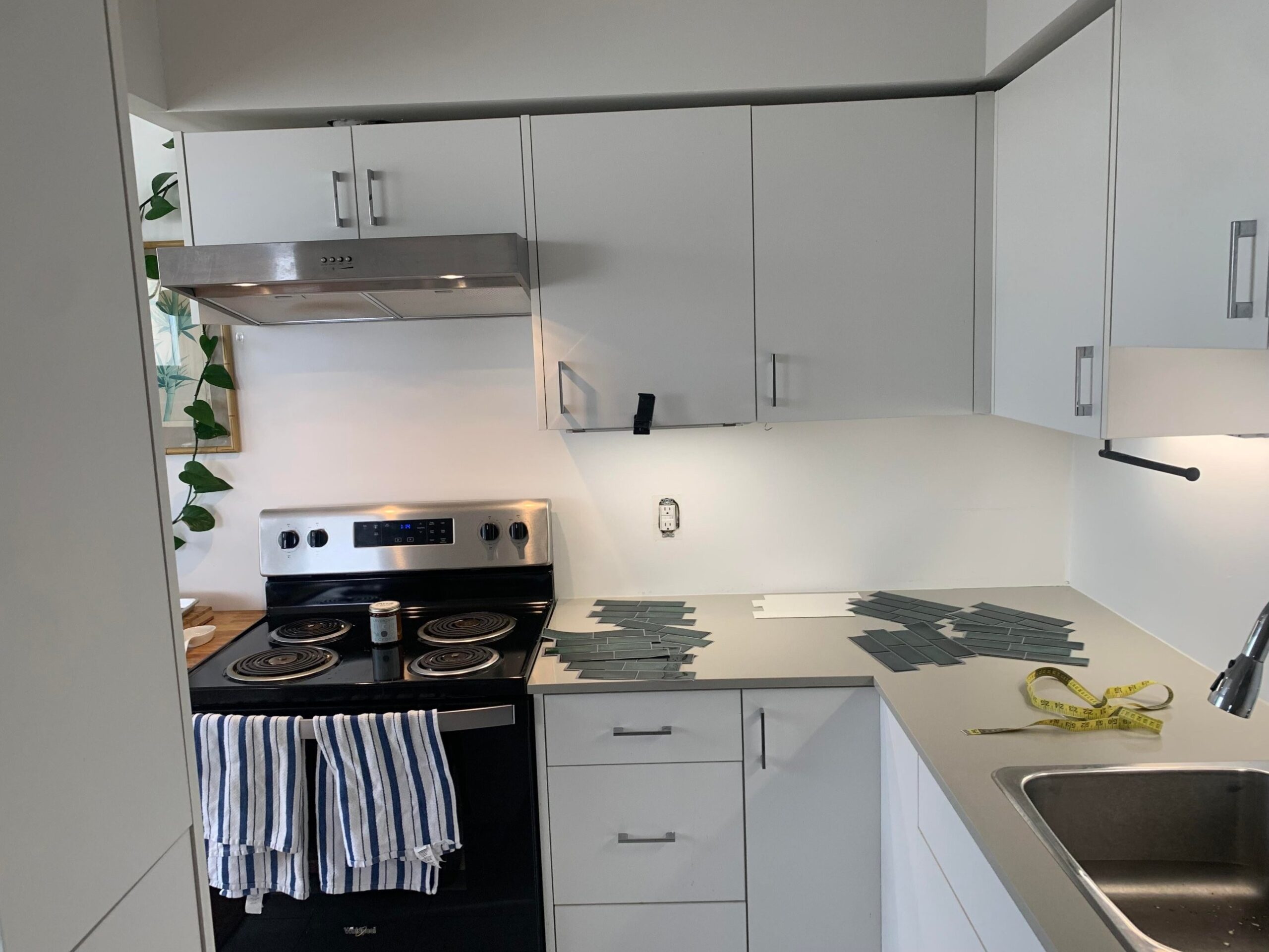Mid-install shot of a rental kitchen stove corner showing a stainless range hood, white cabinets, a bare white backsplash, and uncut green peel-and-stick subway tiles spread across the quartz counter next to the stove.