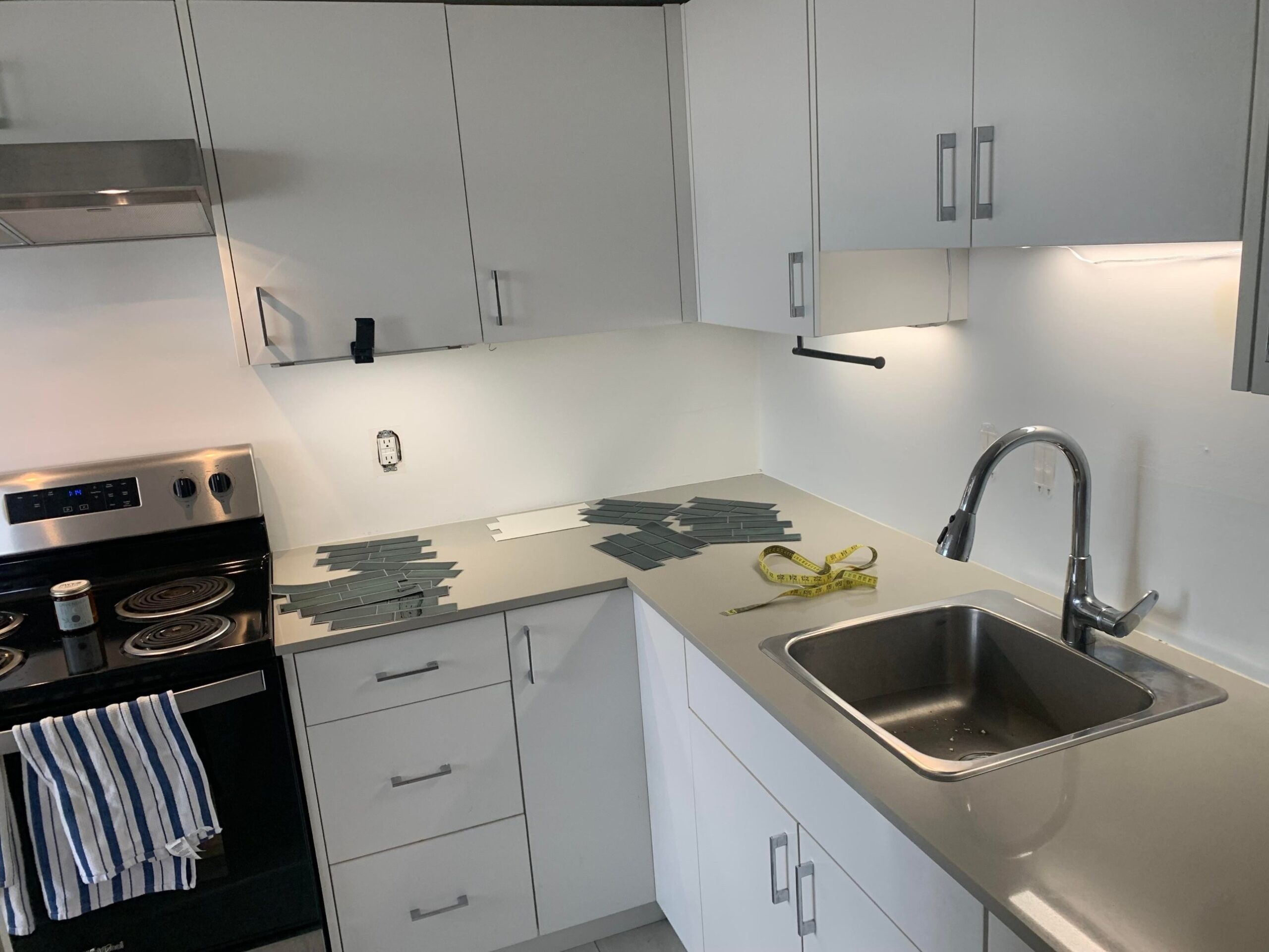 Wide view of a small rental kitchen with white cabinets, a black electric coil stove on the left, a sink on the right, and loose green peel-and-stick subway tiles scattered across the gray quartz counters during install.