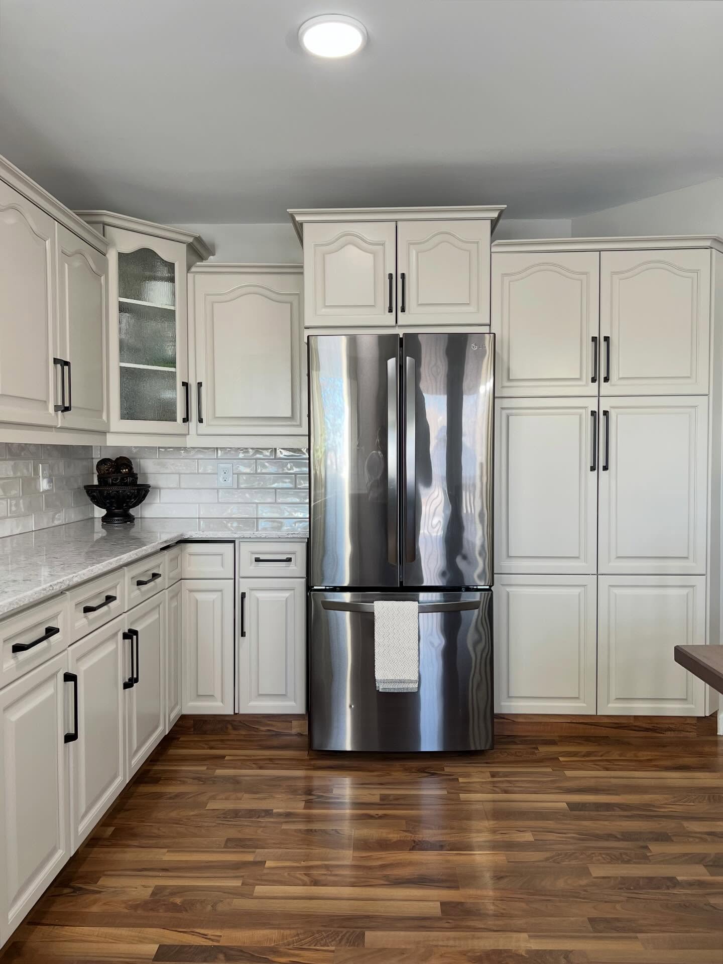 Painted greige kitchen cabinets around a stainless LG French door fridge, with a tall pantry column on the right, a glass-front display cabinet on the left, and warm walnut-tone laminate floors.