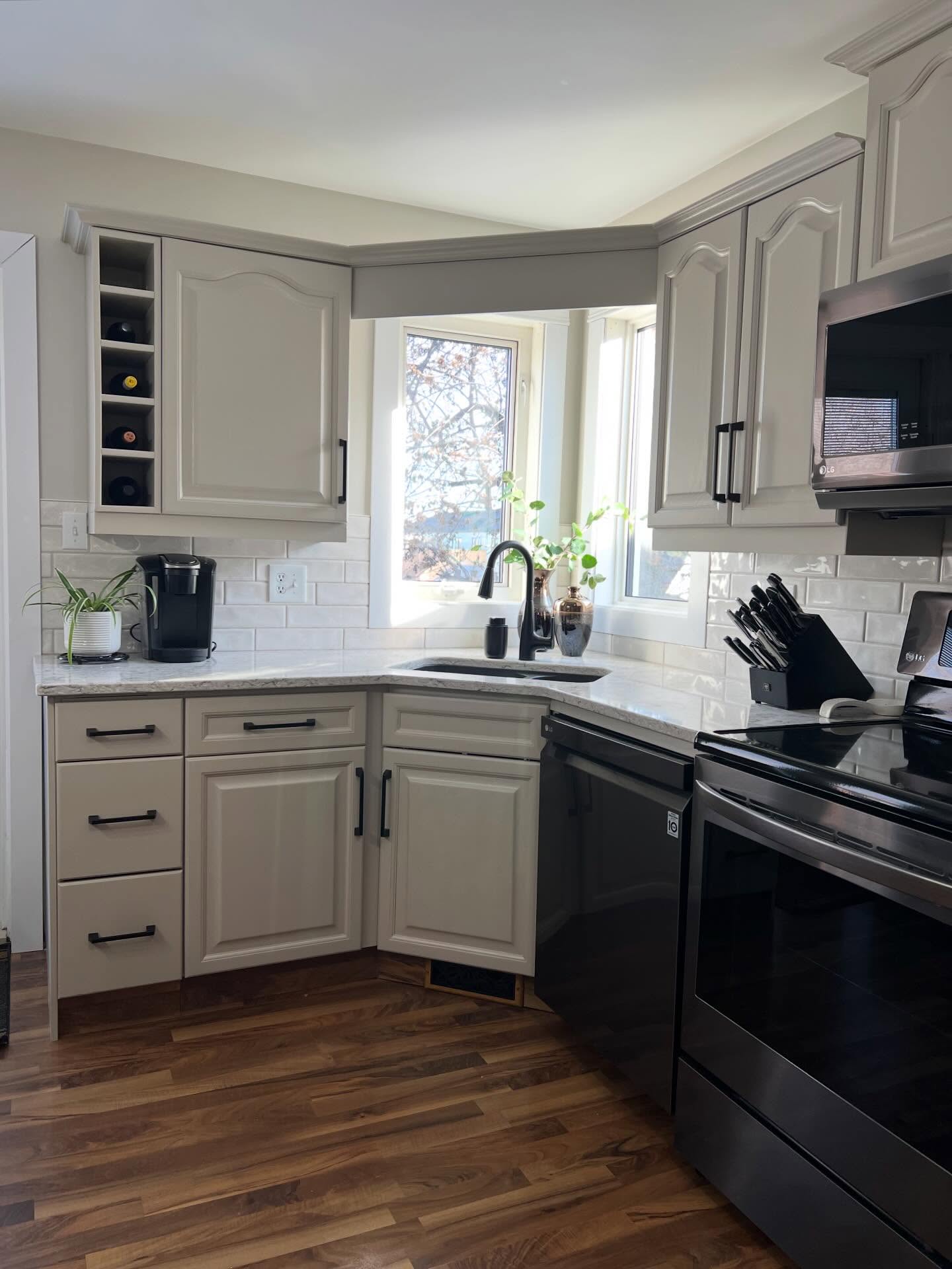 Corner window bay in the painted kitchen with a cream quartz counter, undermount sink, matte black gooseneck faucet, Keurig on the left, and a small open wine cubby built into the upper cabinet.