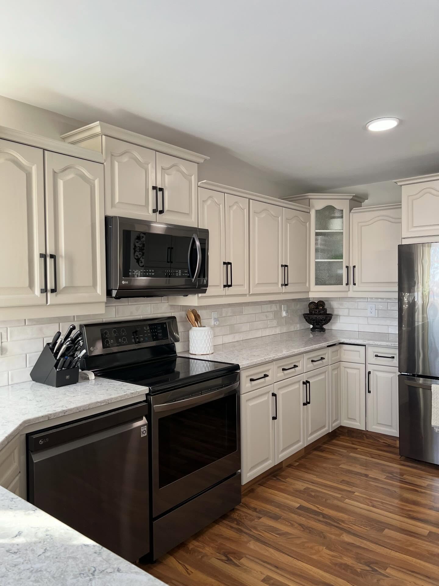 Angle view of the painted kitchen from the island side, showing the stove wall, the window-bay sink, the pantry cabinet, and the fridge all tied together by soft greige cabinets and warm walnut floors.