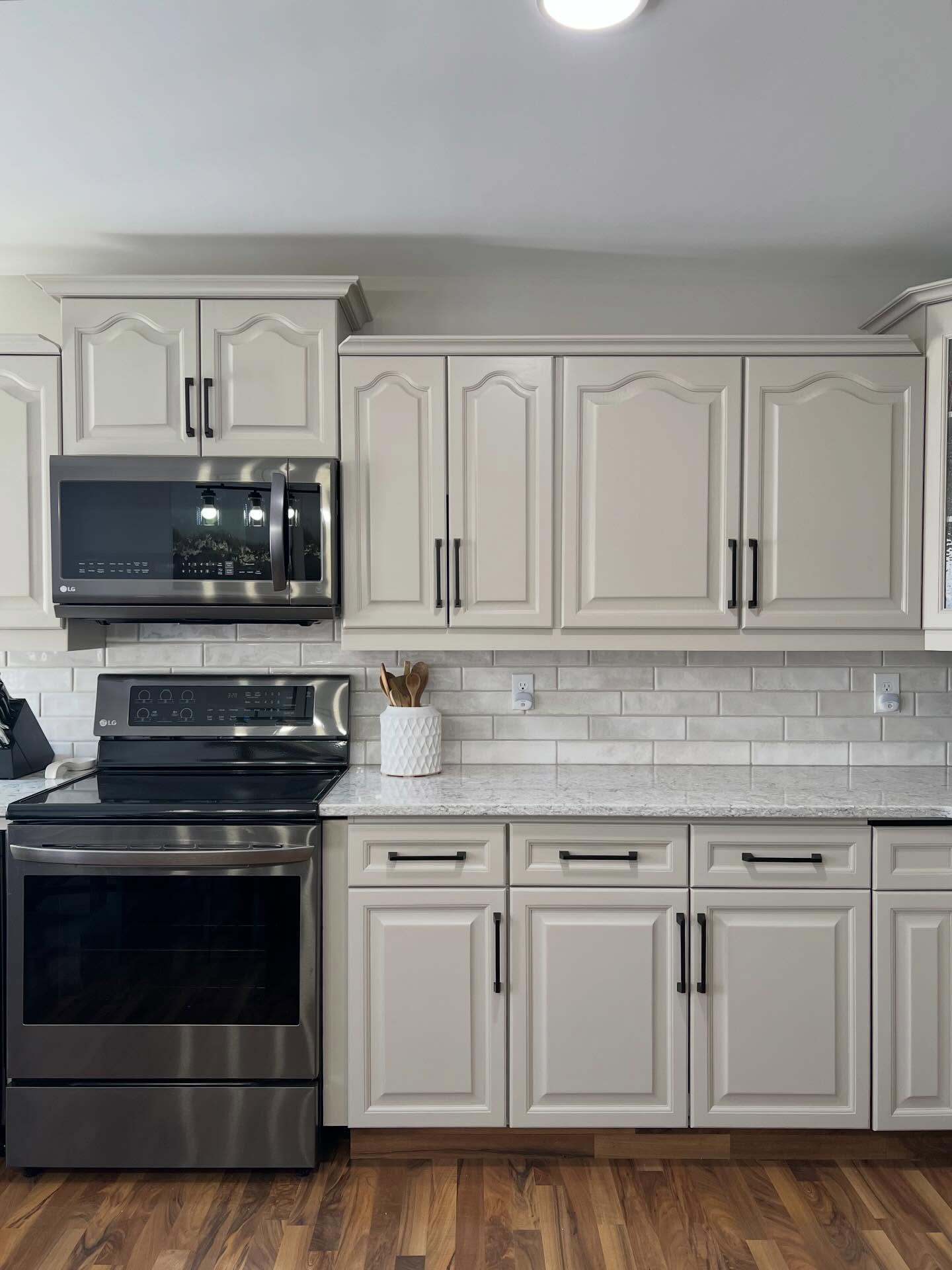 Painted greige kitchen cabinets with cathedral arch upper doors, long matte black bar pulls, cream quartz counters, and a soft off-white brick backsplash around a black stainless LG range and over-range microwave.