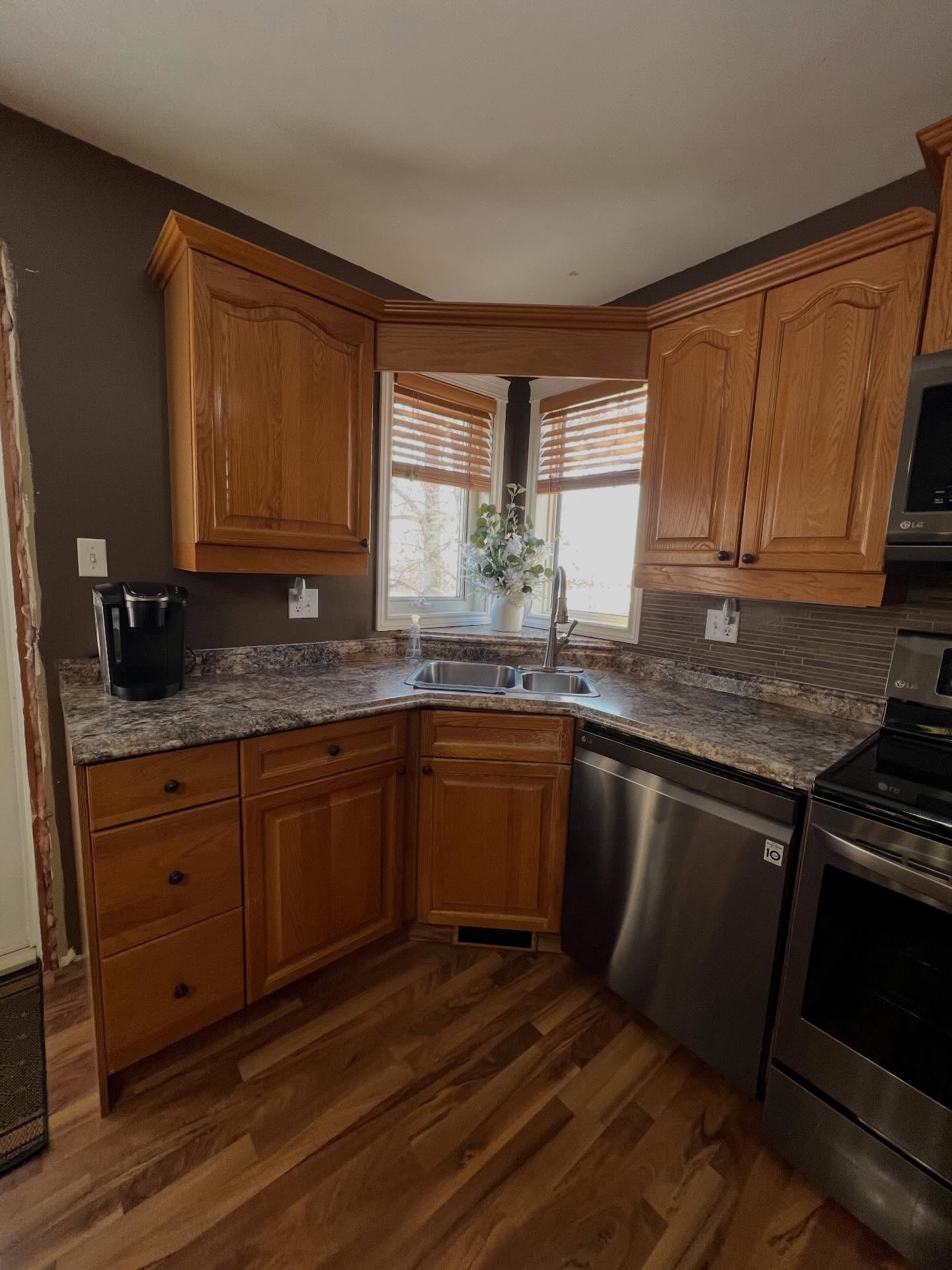 Wide view of the finished kitchen from the dining side, showing all the painted greige arched cabinets, cream quartz counters, soft off-white brick backsplash, matte black hardware, and warm walnut floors tied together.