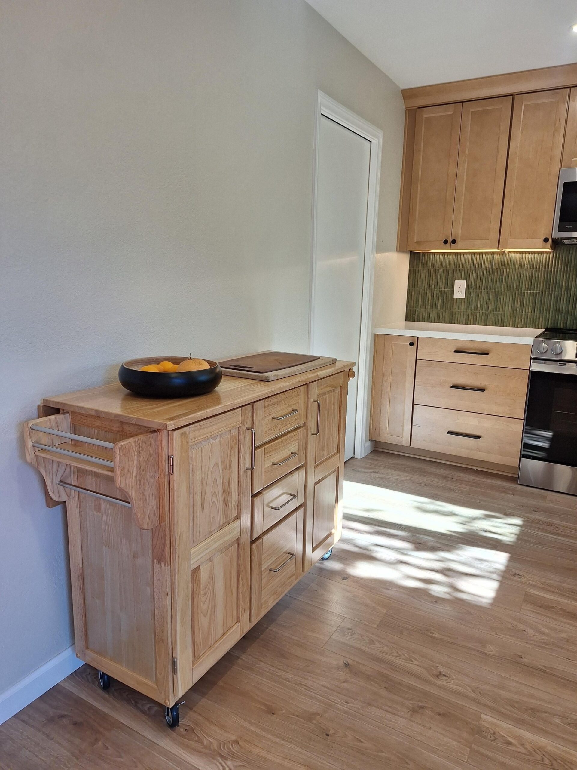 Light natural wood rolling kitchen island with a butcher block top on casters, a towel bar on the side, a black bowl of lemons and a wooden cutting board on top, and a white pocket door closed flush into the wall across the room.