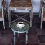 Pair of rustic hand-painted wood armchairs with whitewashed finishes, carved daisy details, and vintage floral toss pillows flanking a small square distressed wood accent table, with a round turned-leg stool painted in weathered sage green and red out front holding a white porcelain cup of black coffee on a saucer with a brass spoon, set over a dark burgundy ceramic tile floor.