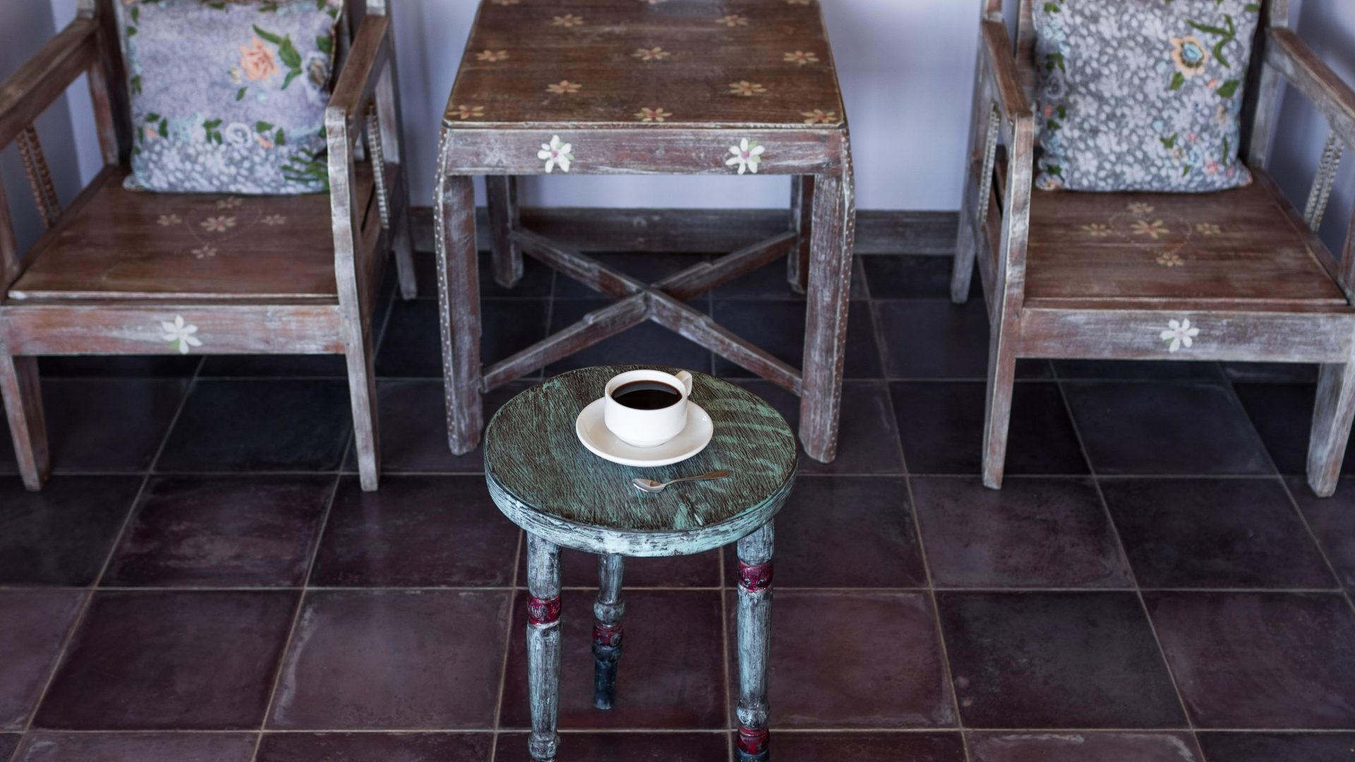 Pair of rustic hand-painted wood armchairs with whitewashed finishes, carved daisy details, and vintage floral toss pillows flanking a small square distressed wood accent table, with a round turned-leg stool painted in weathered sage green and red out front holding a white porcelain cup of black coffee on a saucer with a brass spoon, set over a dark burgundy ceramic tile floor.