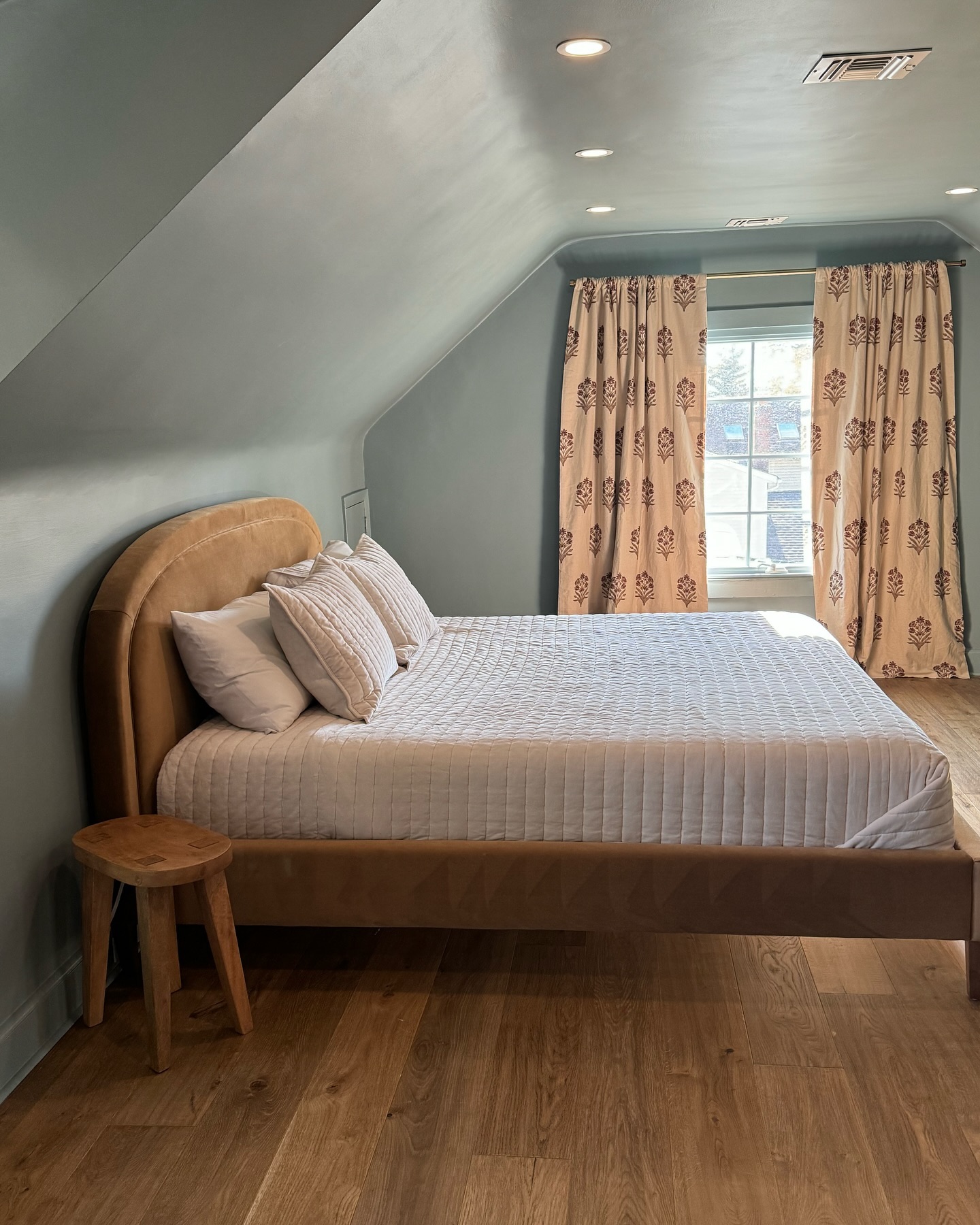 Modern attic bedroom with sage blue painted walls and sloped ceiling, tan velvet arched platform bed, quilted bedding, cream block print curtains, and wide plank oak floors