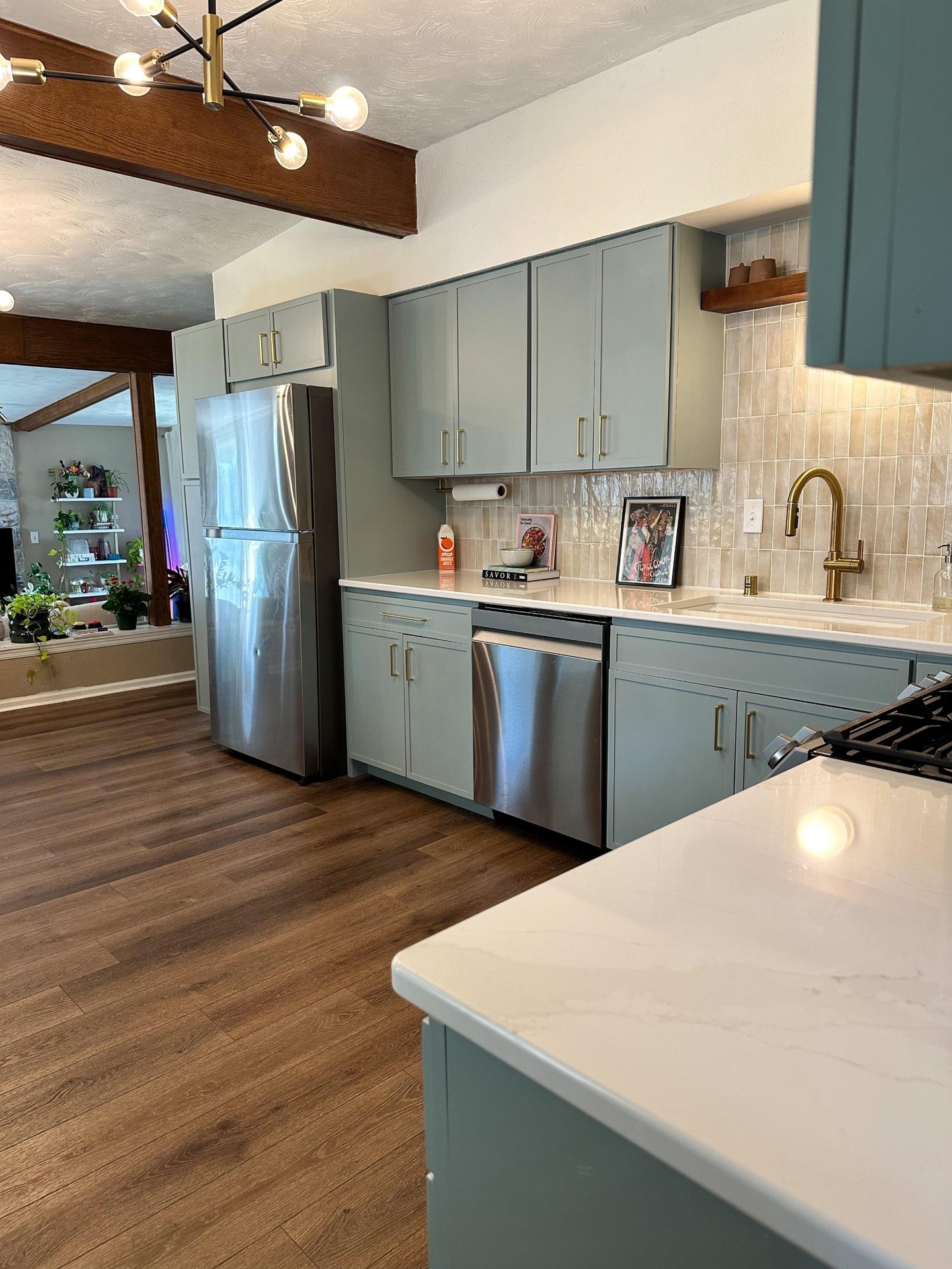 Close view of the finished kitchen showing a white quartz countertop with marble veining, sage blue cabinets, vertical beige tile backsplash, a brushed brass pull-down faucet, an open warm wood floating shelf and a small framed vintage French poster leaning against the backsplash.
