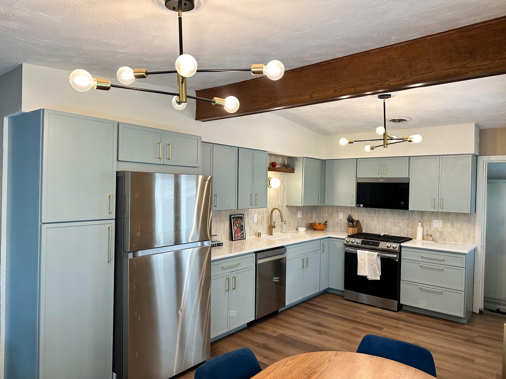 Finished kitchen with soft sage blue shaker cabinets, white quartz counters, vertical beige subway tile backsplash, brushed brass pulls, a stainless gas range and fridge, and black and brass sputnik chandeliers overhead.