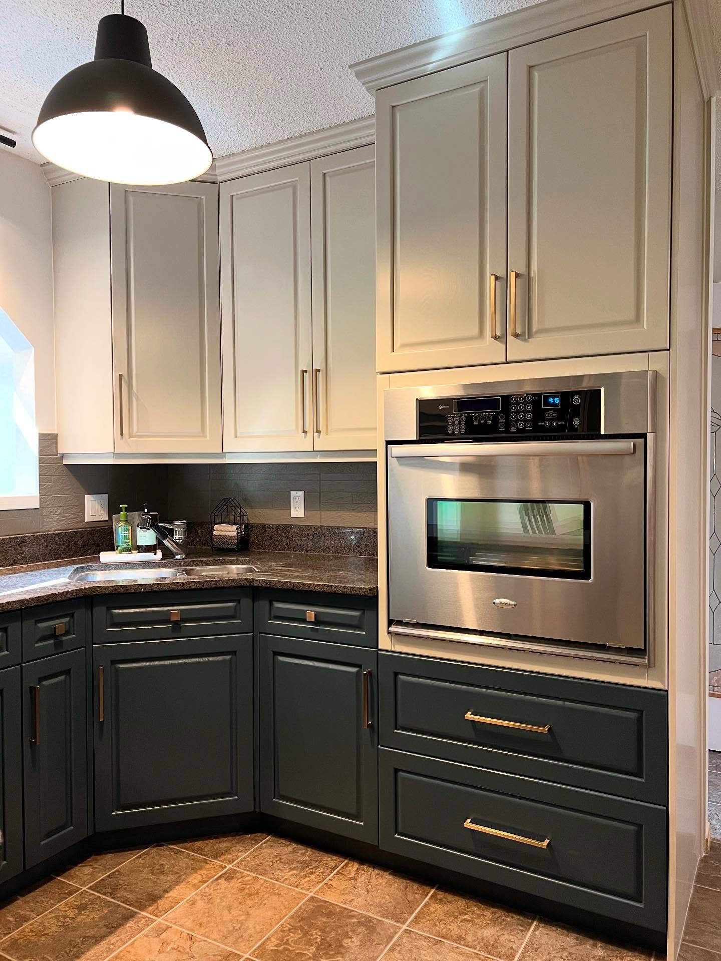 Painted two-tone kitchen sink corner with Hazy Skies sage upper cabinets, Lead Grey dark forest green lower cabinets with long brass bar pulls, a stainless Whirlpool wall oven, a matte black industrial pendant, and a painted gray stone-look backsplash.