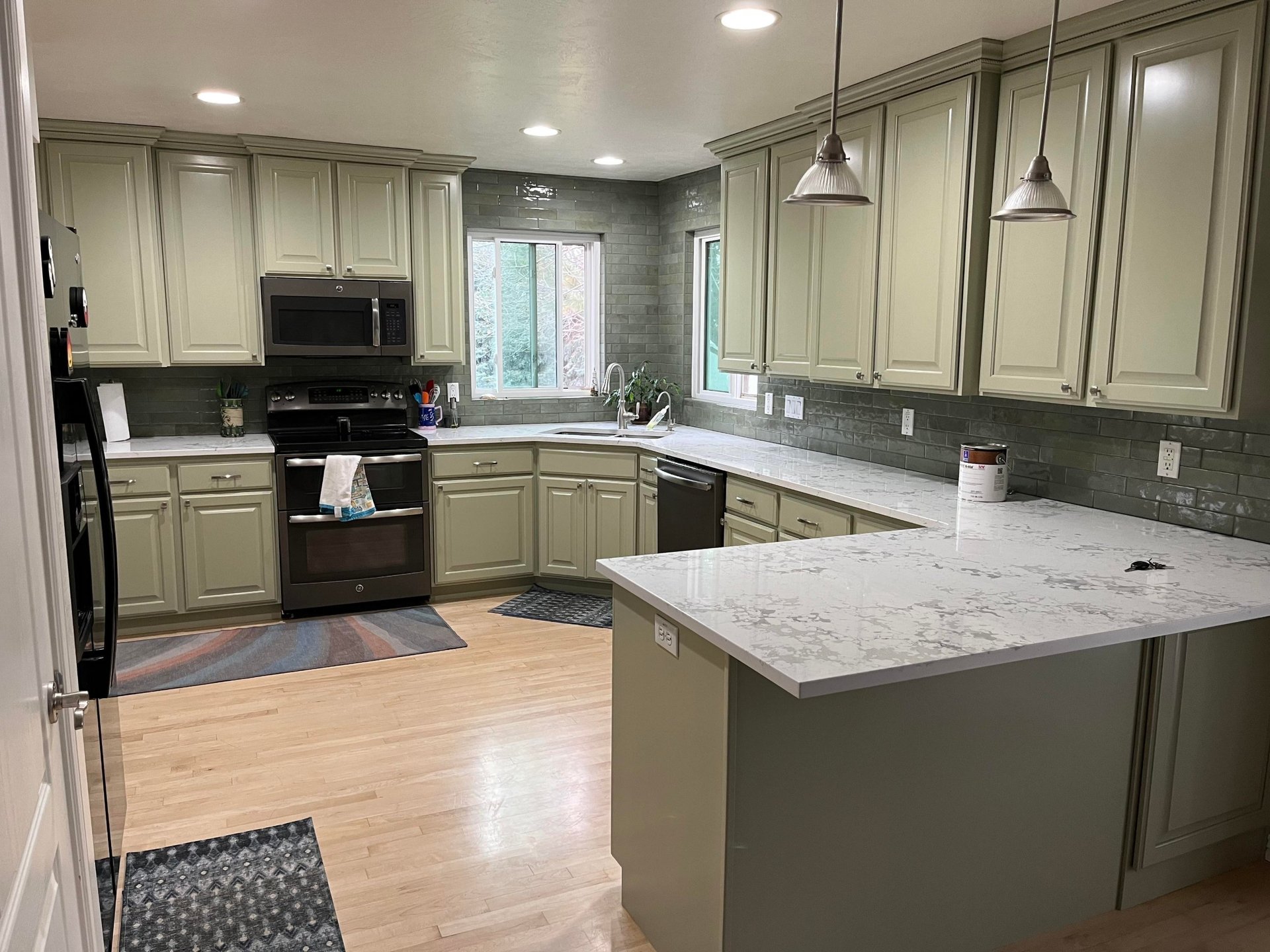 Full view of a G-shaped kitchen with sage green painted cabinets, dark sage glossy subway tile running to the ceiling and wrapping the windows, white quartz countertops with heavy gray veining, stainless undermount sink, and black GE range.