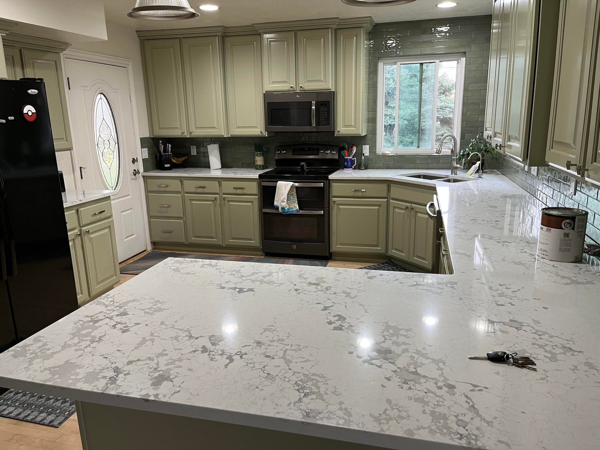 Sage green painted kitchen cabinets with a large white and gray veined quartz peninsula, dark sage glossy subway tile backsplash, black appliances, undermount stainless sink, and silver pendant lights.
