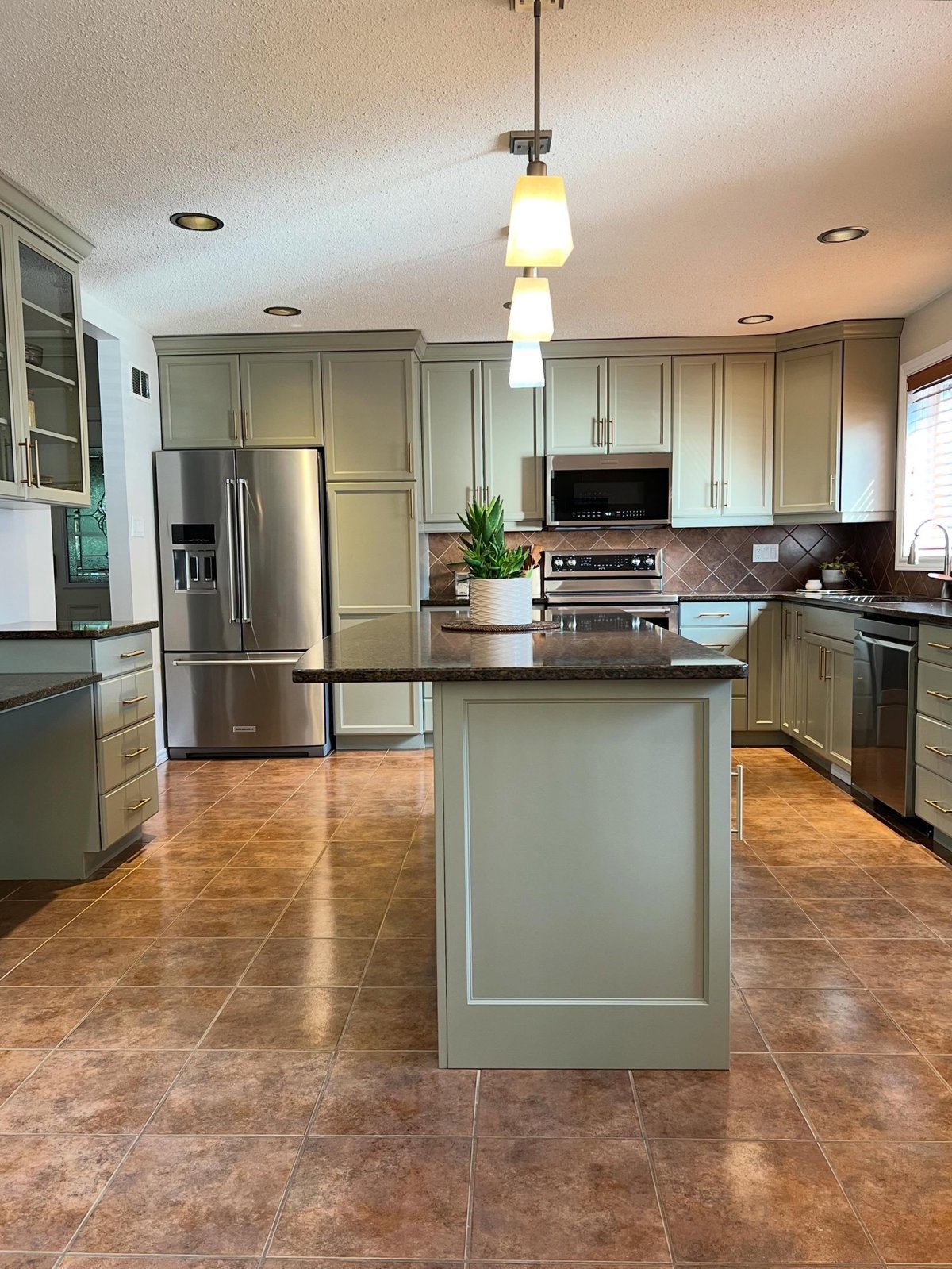 Same kitchen after painting, with sage green Honed Soapstone cabinets, brass bar pulls, Pale Oak walls, the same brown granite island, brown diamond backsplash and terracotta floor tile.
