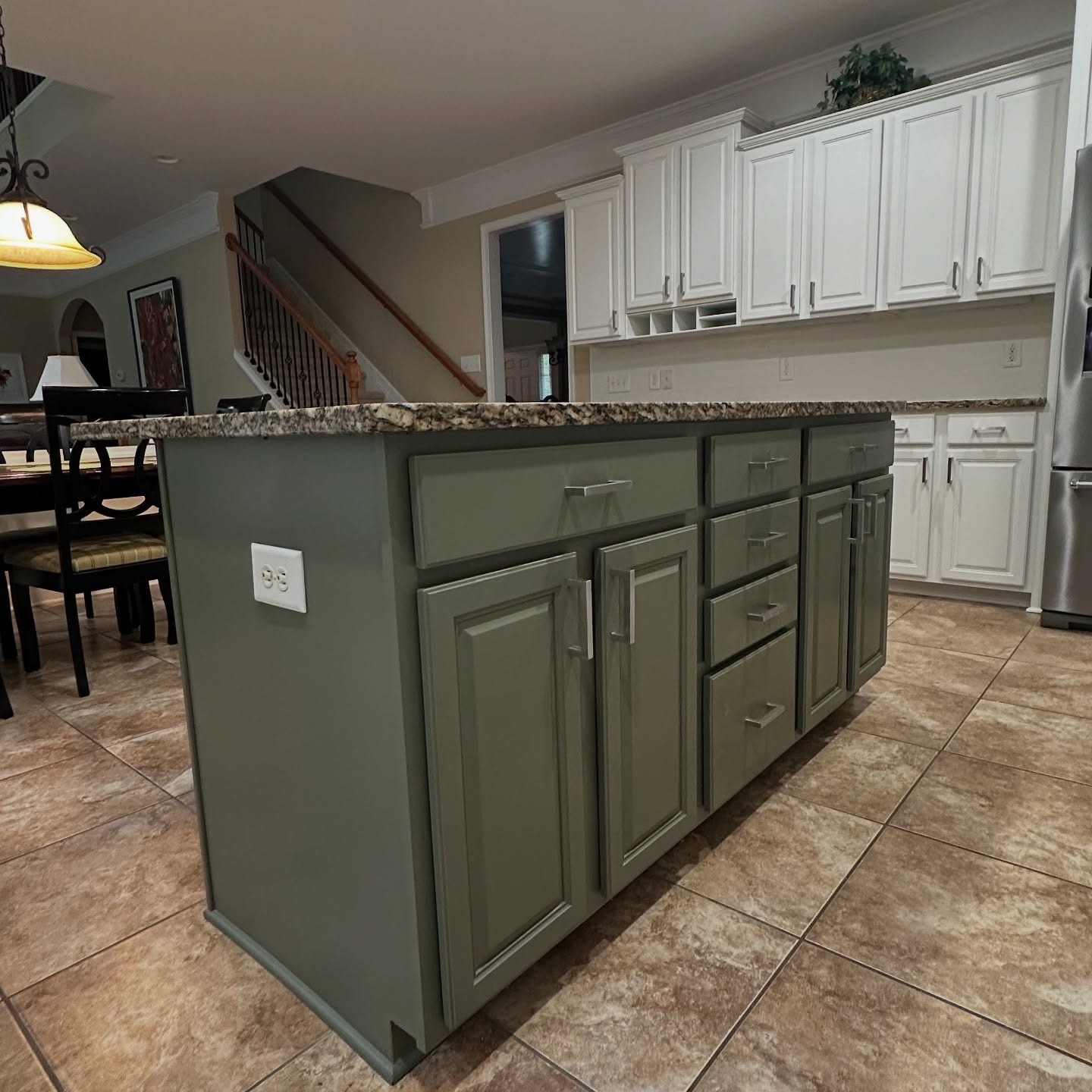 Side view of a kitchen island painted muted sage green with silver bar pulls, a cream and brown granite top, white perimeter cabinets in the background, and warm porcelain floor tile.