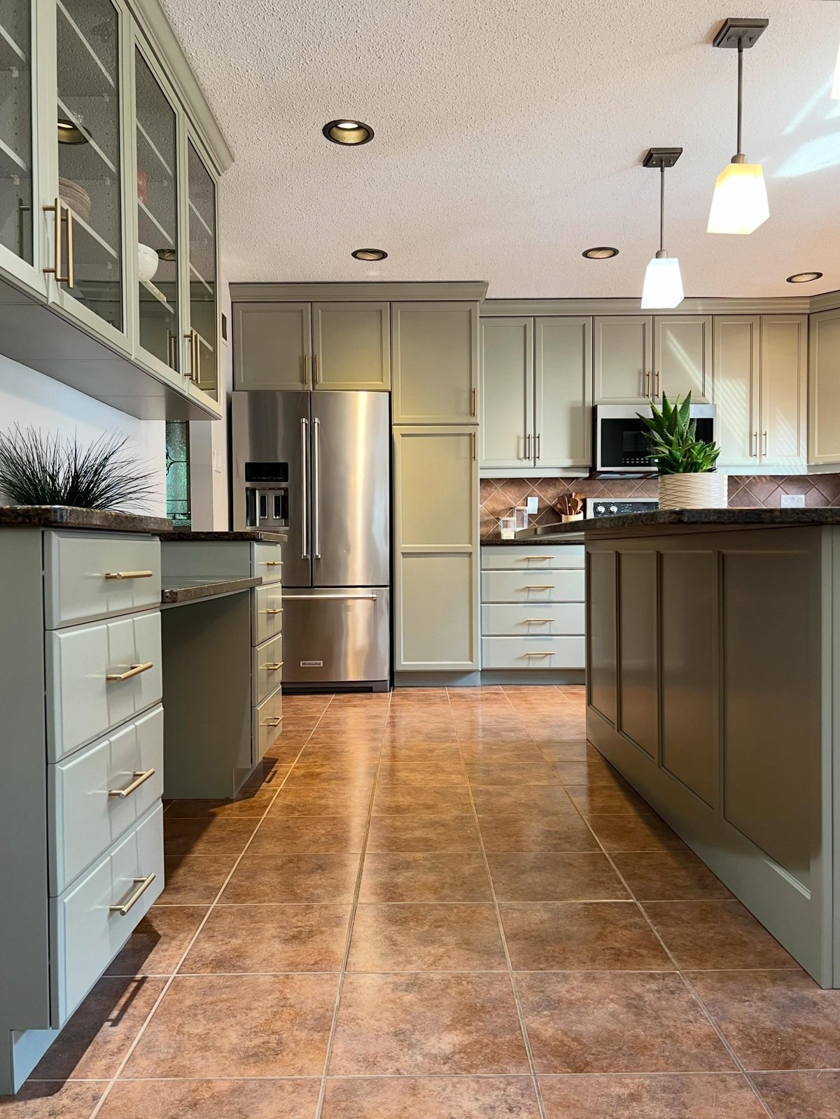 Wide view of the finished kitchen with sage green Honed Soapstone cabinets, brass bar pulls, Pale Oak walls, a center island with brown granite top, three pendant lights, stainless steel fridge and the original brown diamond backsplash and terracotta floor.