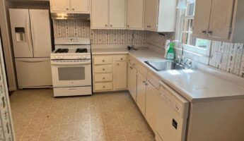Dated 1990s kitchen with white raised-panel cabinets, floral border wallpaper, beige patterned vinyl tile floor, a white gas range, a white French-door fridge at the end of the counter run, and a stainless drop-in sink under a window.