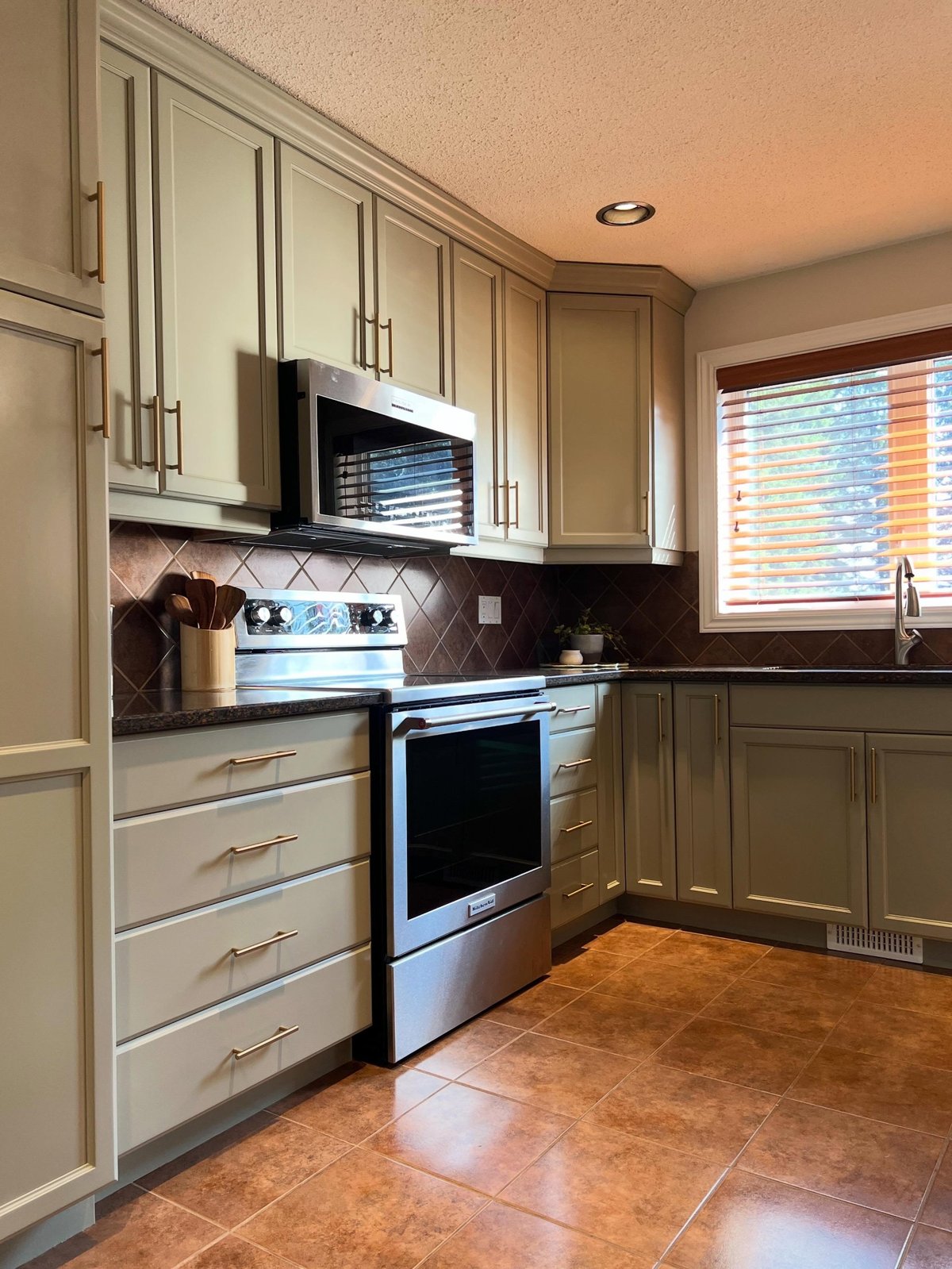 Close-up of the same stove and window wall after painting, sage green Honed Soapstone cabinets with brass bar pulls, white trim around the window, the same brown diamond backsplash and granite counters.