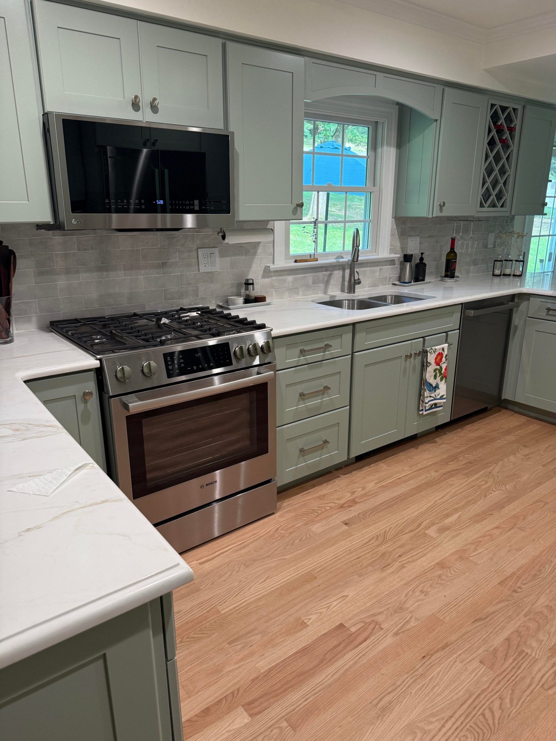 Wide view of the finished kitchen with sage green shaker cabinets, white quartz counters with gold veining, a gray brick tile backsplash, a stainless gas range, and a matte red oak hardwood floor.
