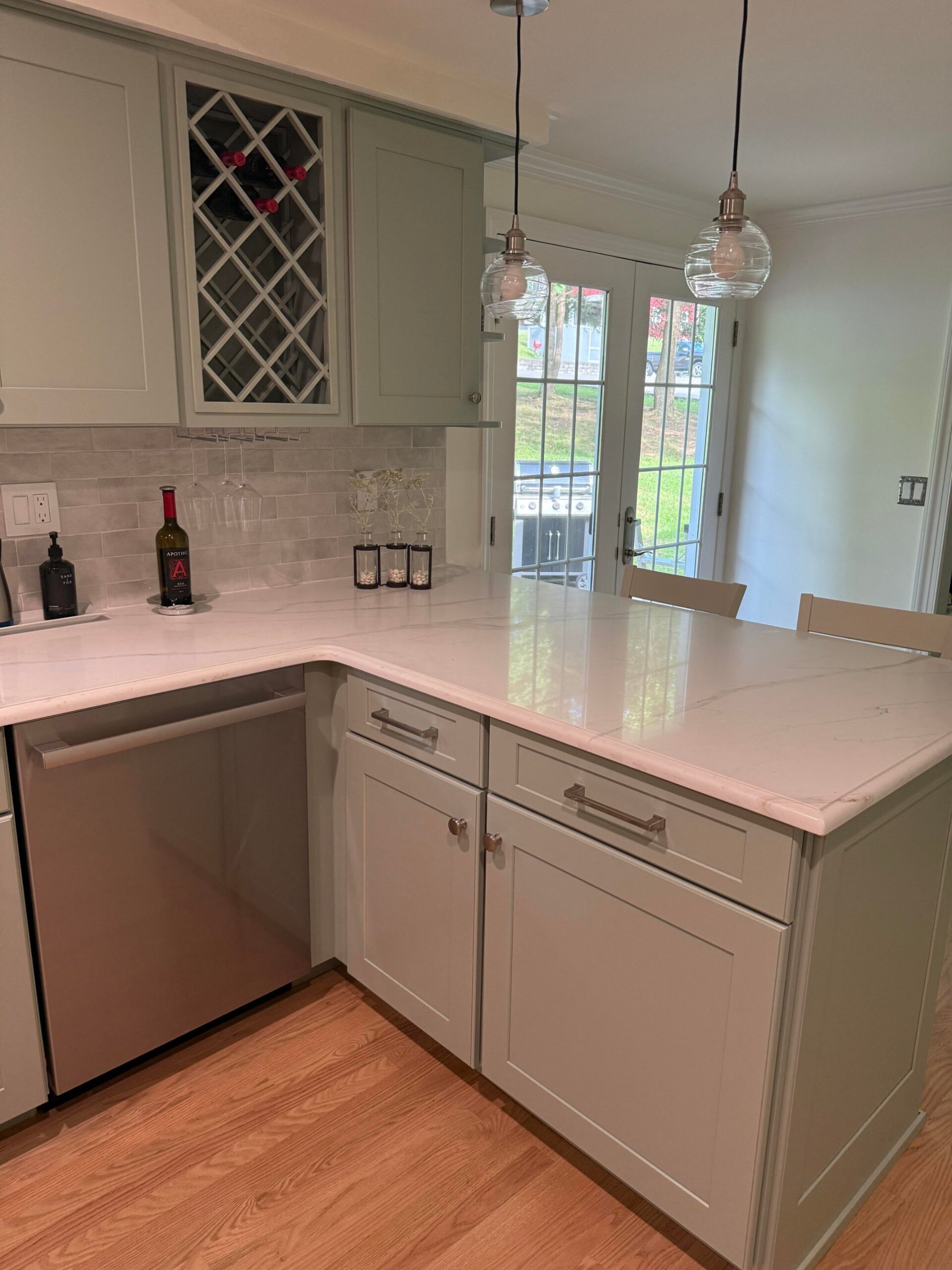 Sage shaker peninsula with white quartz counter, two glass pendant lights above, a diamond-pattern wine lattice cabinet, and French doors leading to the backyard.