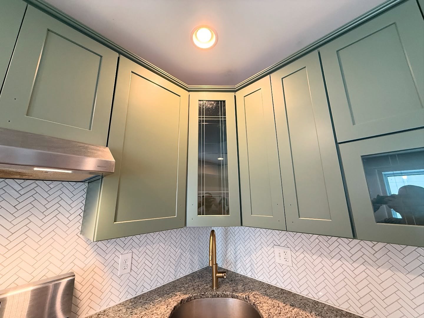 Close view of corner sink cabinets painted in sage green Rosepine with brass gooseneck faucet, speckled granite counter, white herringbone backsplash, and leaded glass cabinet detail.