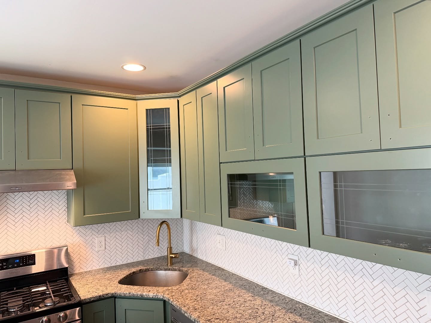 Close-up of upper sage green cabinets in Rosepine showing leaded glass panels, white herringbone backsplash, brass faucet, and crown molding detail.