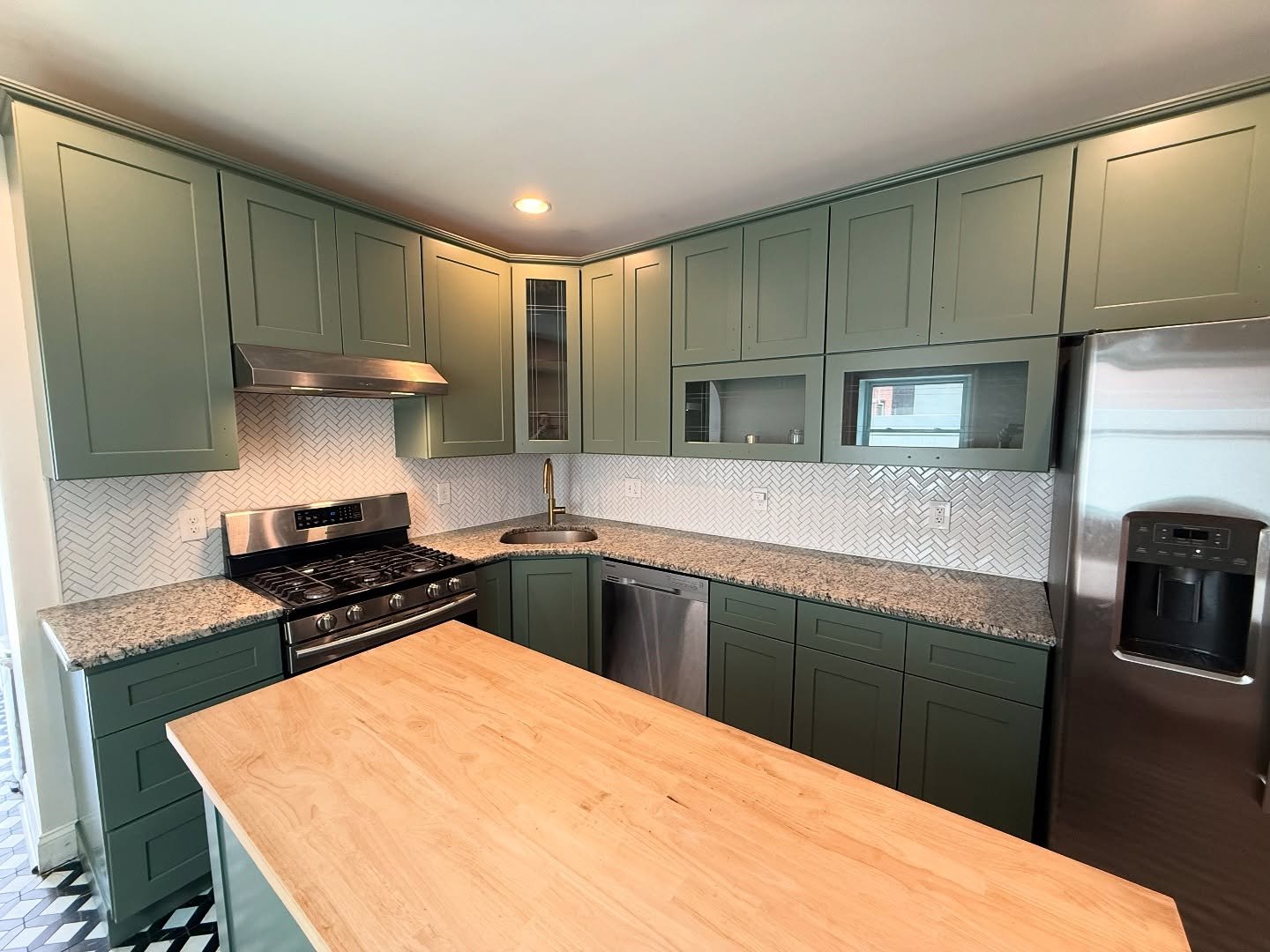 Soft sage green shaker kitchen in Benjamin Moore Rosepine with speckled granite counters, white herringbone backsplash, brass faucet, stainless appliances, and butcher block island.