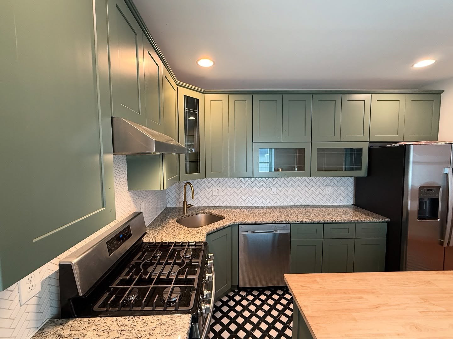 Sage green Rosepine kitchen with stainless range, brass faucet over corner sink, white herringbone backsplash, speckled granite counters, and butcher block island edge.